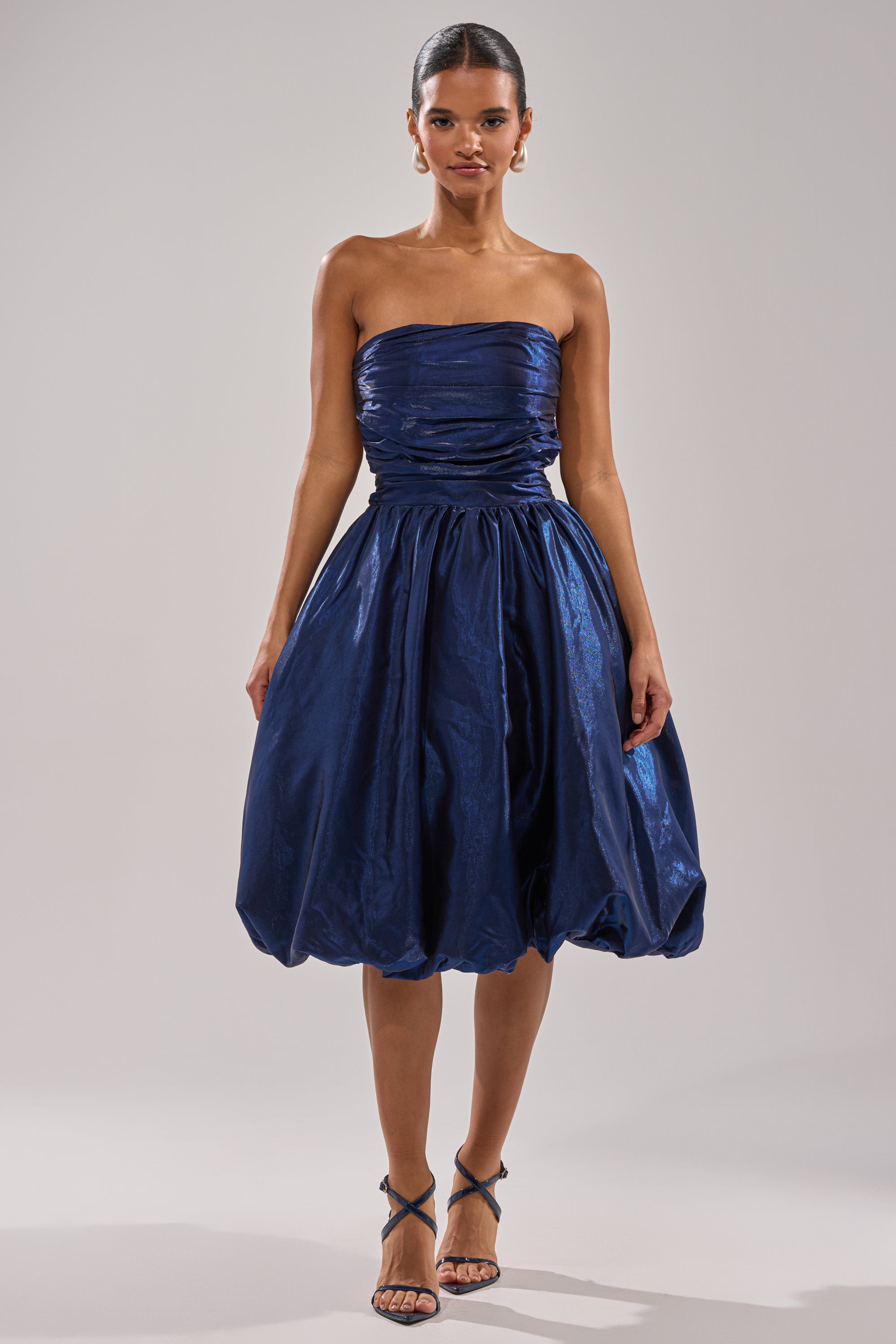 A woman stands against a simple gray background wearing the LUXE LIFE SATIN MIDI DRESS, a strapless dark blue midi with ruched details and a voluminous bubble skirt, paired with strappy high-heeled sandals. 6