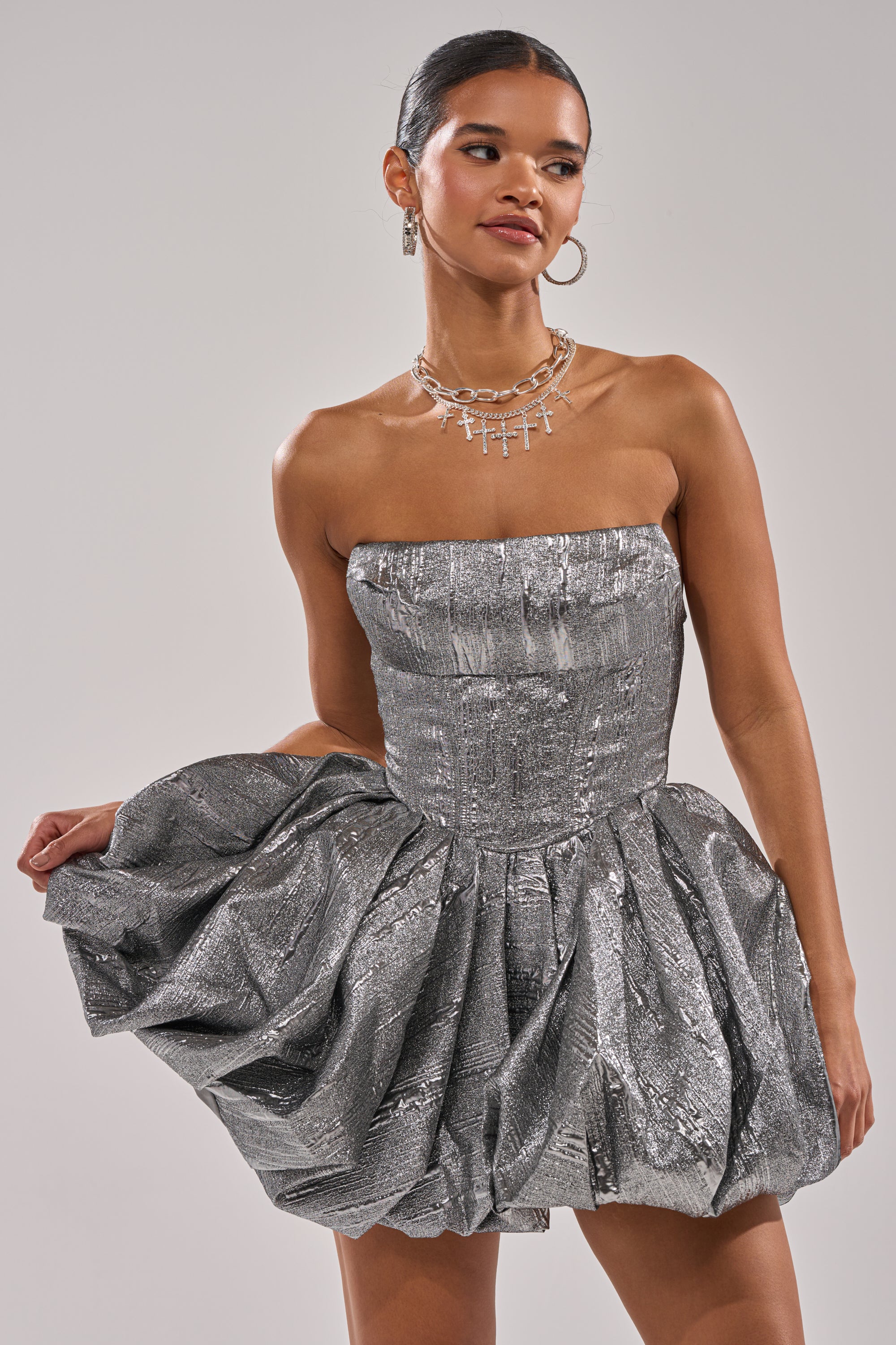 A woman models the STAR OF THE NIGHT SILVER MINI DRESS with a strapless design and a voluminous bubble skirt, paired with hoop earrings and layered necklaces, her hair pulled back against a plain light background.