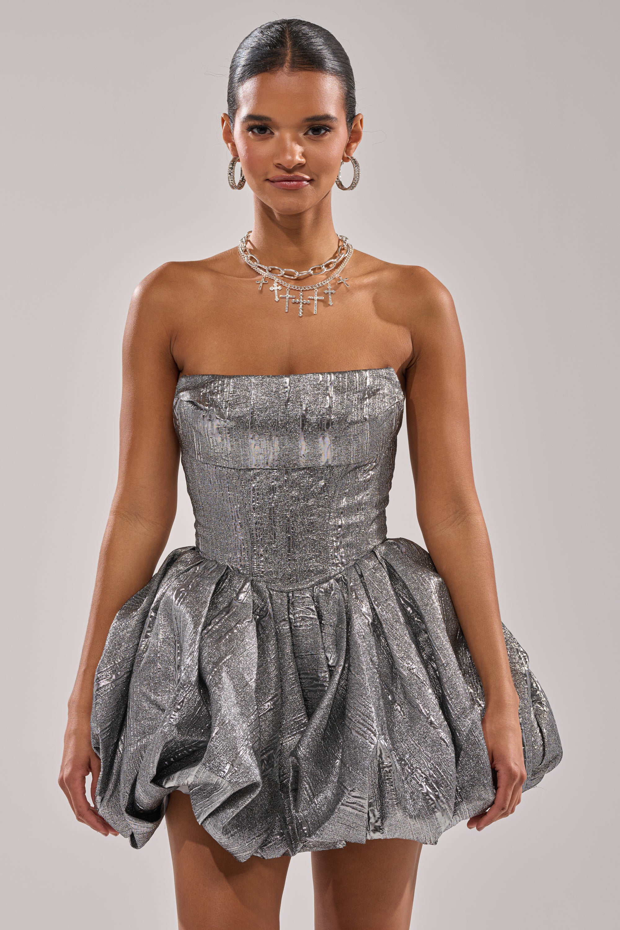A woman poses against a light gray background wearing the STAR OF THE NIGHT SILVER MINI DRESS, featuring a strapless corset top and textured bubble skirt, styled with silver hoop earrings and layered necklaces.