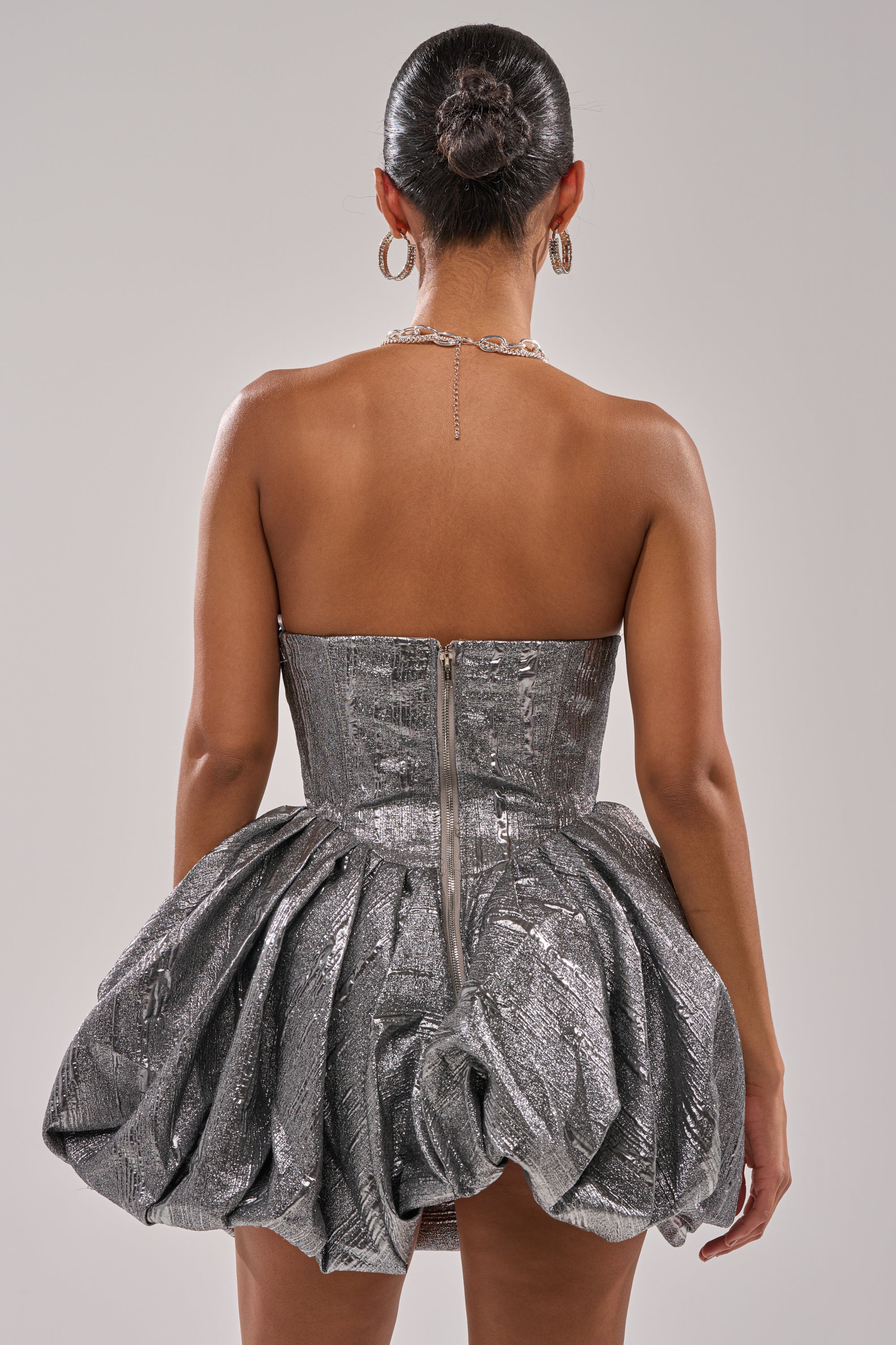 A woman with her hair in a sleek bun is shown from behind, wearing the STAR OF THE NIGHT SILVER MINI DRESS—a strapless mini with a voluminous bubble skirt and visible back zipper—accessorized with hoop earrings and a delicate necklace.