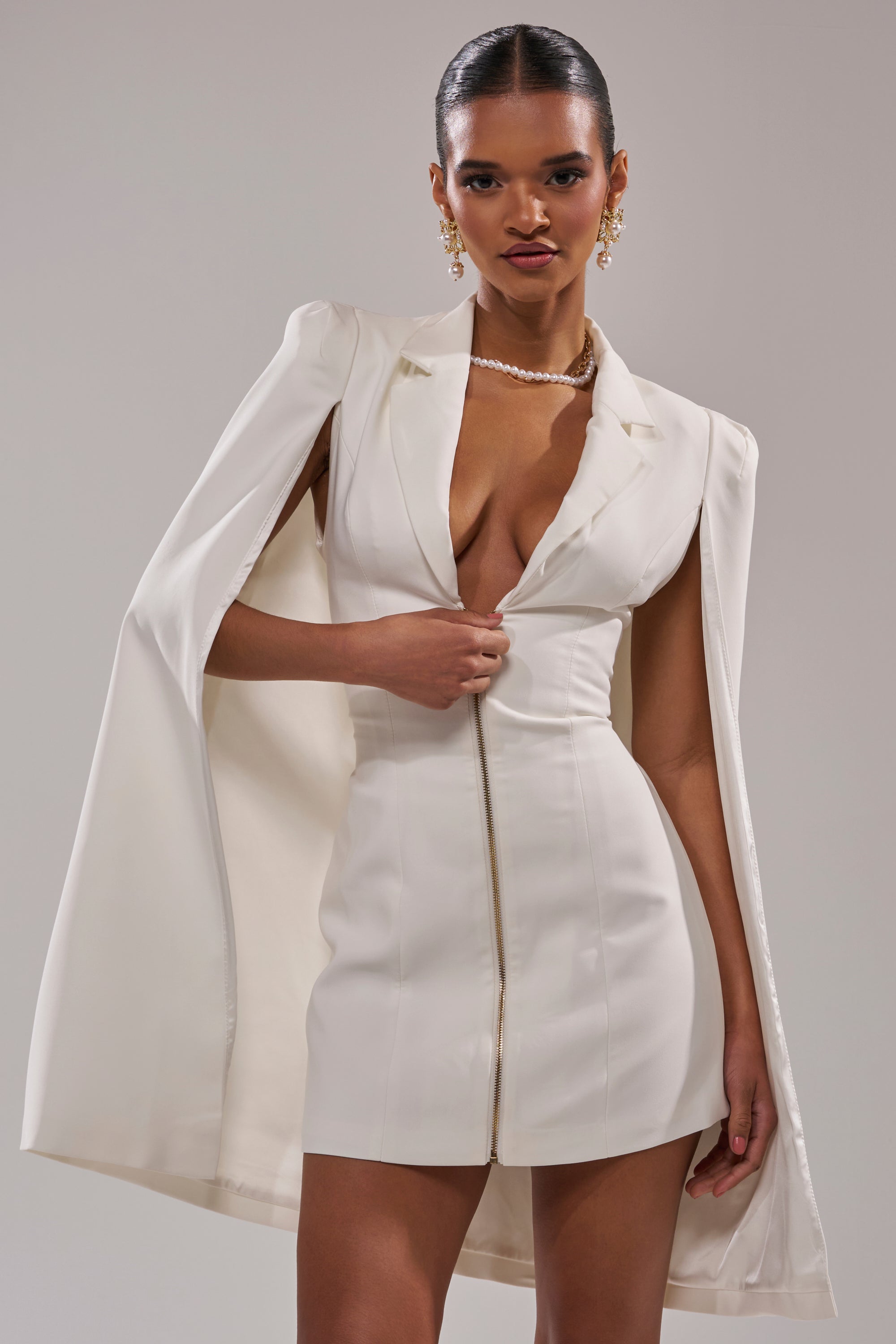 A woman stands confidently in the STRICTLY BUSINESS BLAZER DRESS, a sleek white suiting piece with a cape-style jacket, deep V-neckline, and gold zipper. She accessorizes with pearl jewelry, gold earrings, and wears her hair in a slicked-back bun. 1