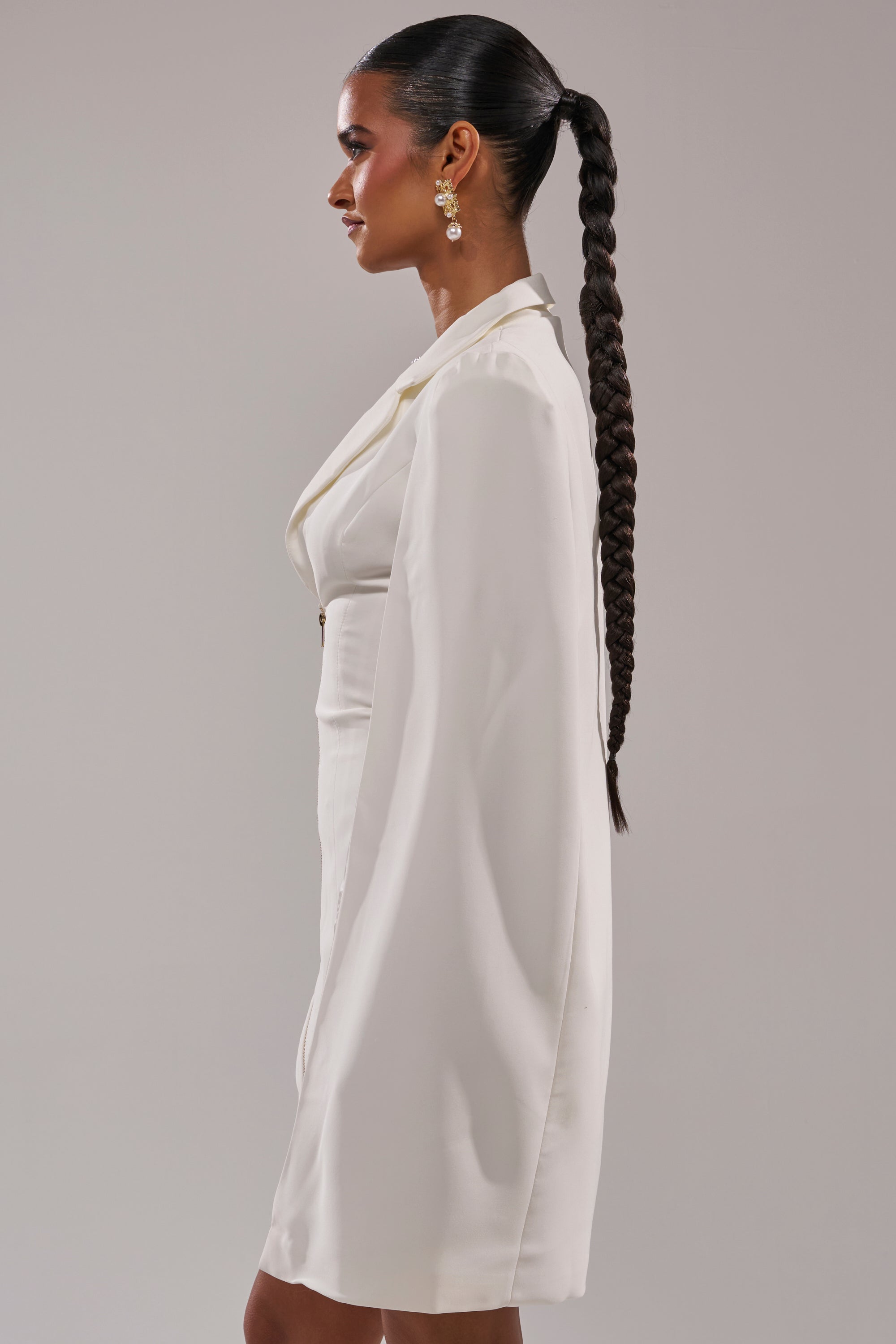 A woman in the STRICTLY BUSINESS BLAZER DRESS with cape sleeves and earrings stands in profile, her hair styled in a long, sleek braid against a plain light background. 3