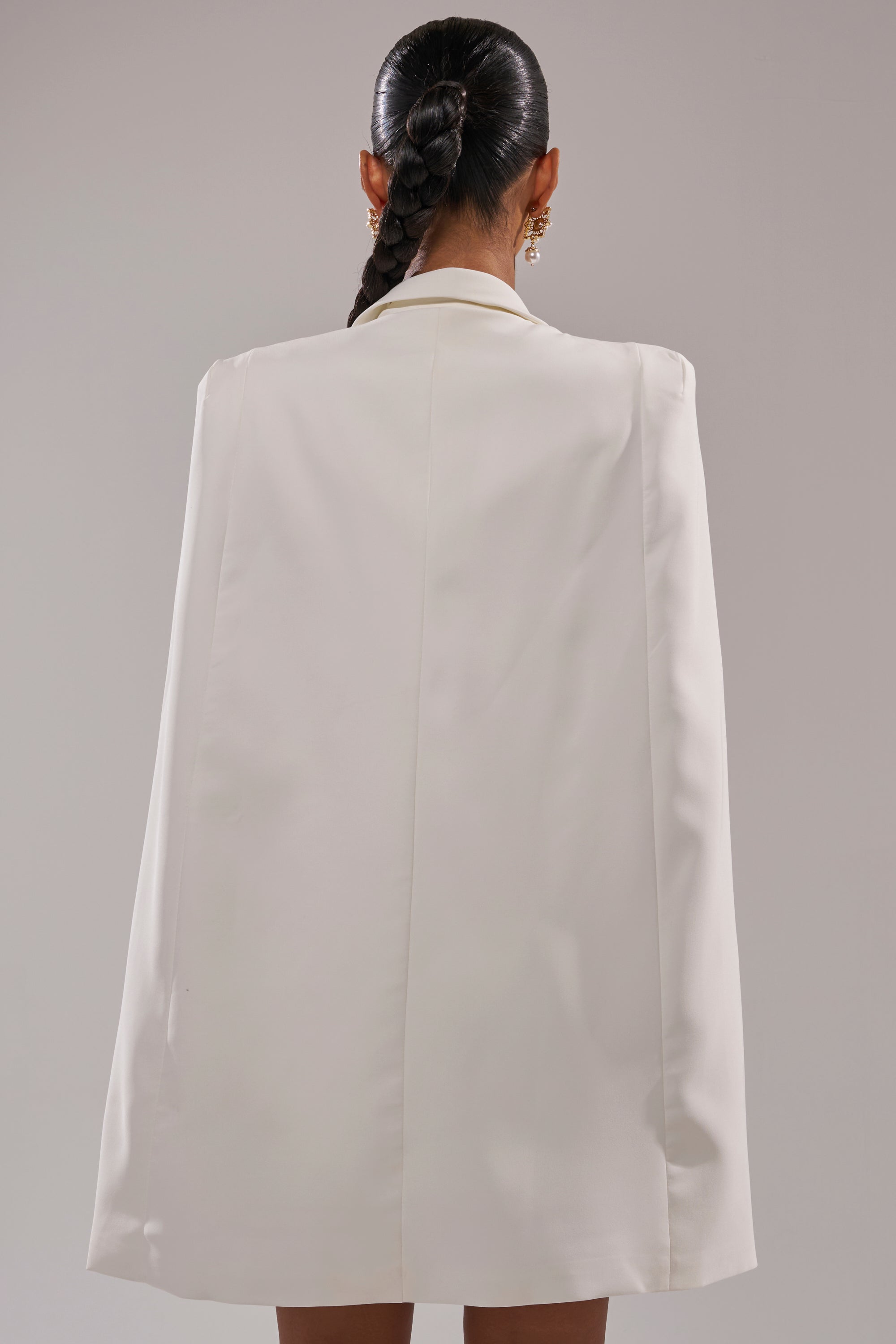 A woman with a long, dark braid faces away, highlighting the STRICTLY BUSINESS BLAZER DRESS. The simple neutral background emphasizes the modern elegance of her tailored white look. 4