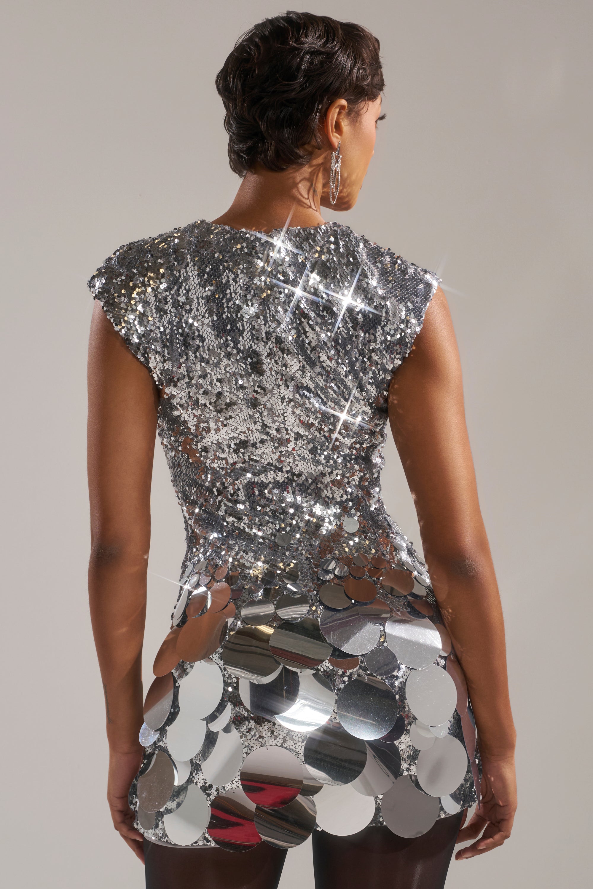 A person with short dark hair is shown from behind in the DANCE THE NIGHT AWAY SEQUIN MINI DRESS IN SILVER, featuring large shiny discs on the lower half—perfect for holiday parties. Hoop earrings complement the sparkling, light-reflecting look.