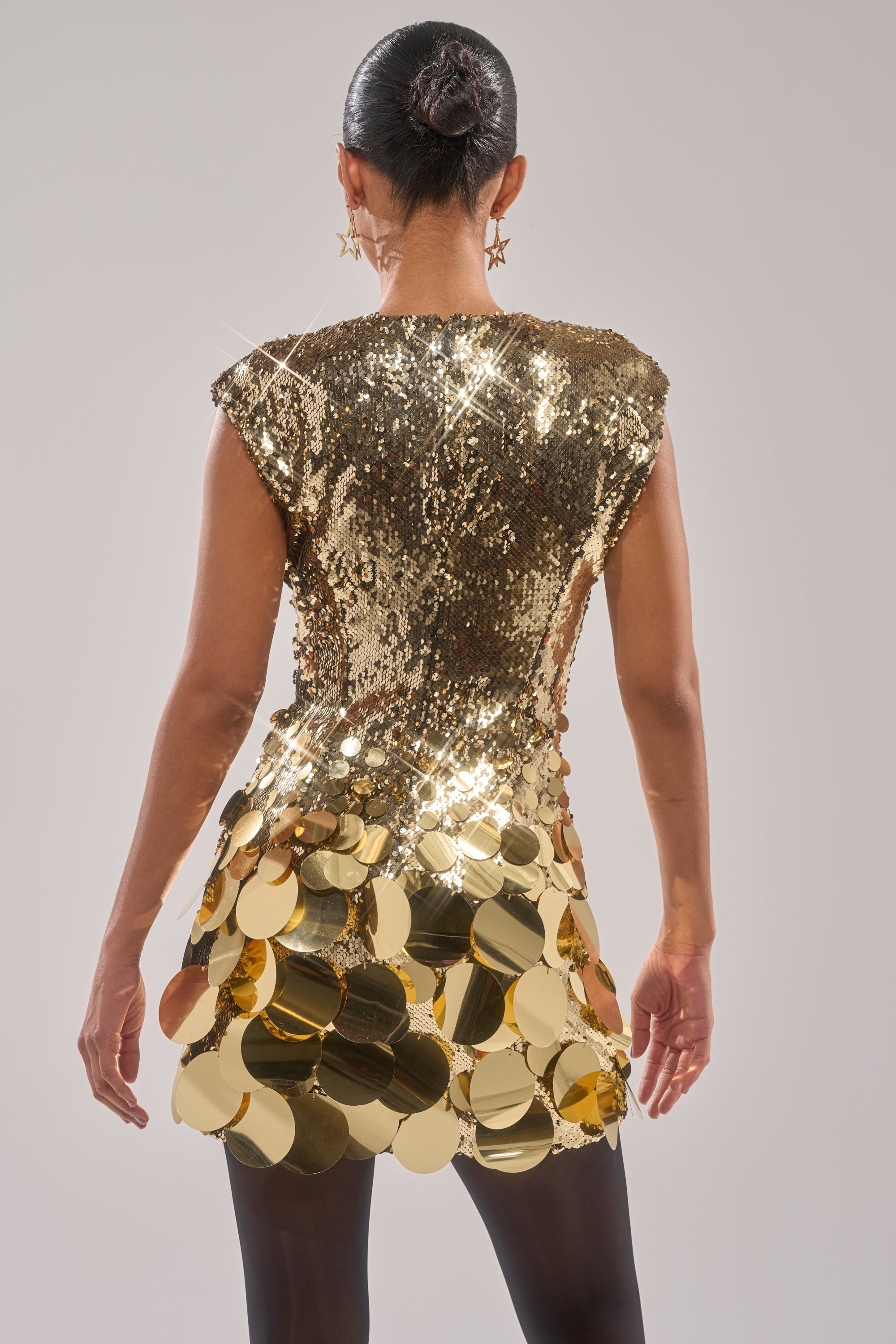 A woman with dark hair in a bun is seen from the back wearing the DANCE THE NIGHT AWAY SEQUIN MINI DRESS IN GOLD, styled with gold star earrings and black tights, against a plain light background.