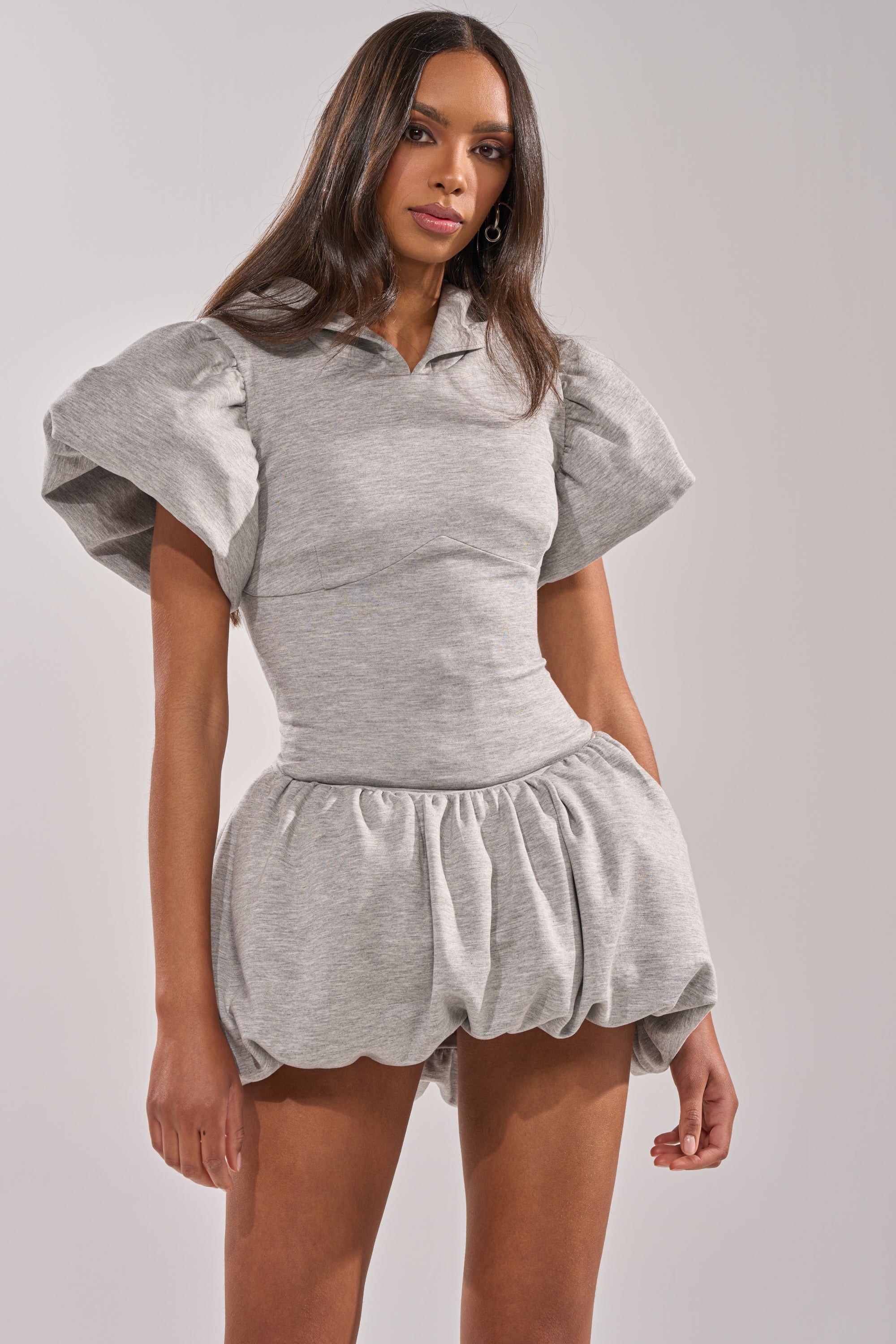 A woman stands against a plain background wearing the GIRL POWER FRENCH TERRY MINI DRESS, a light gray piece with a sculptural silhouette, puffed short sleeves, and a bubble-style skirt. 2