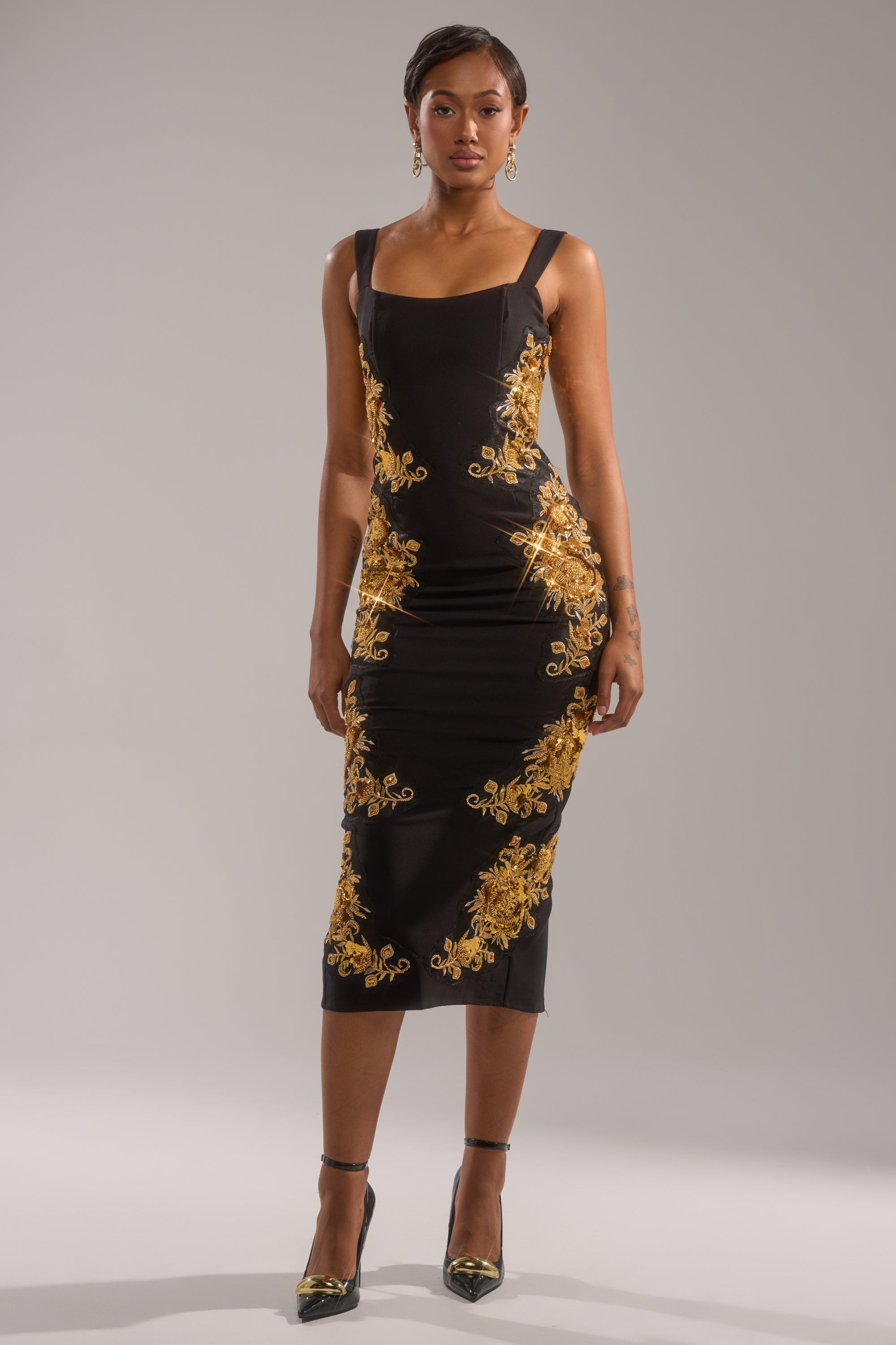 A woman wears the PARISIAN FLORAL EMBROIDERED MAXI DRESS—a fitted, sleeveless black dress with ornate gold embroidery—paired with black heels and gold earrings, standing against a plain light gray background.