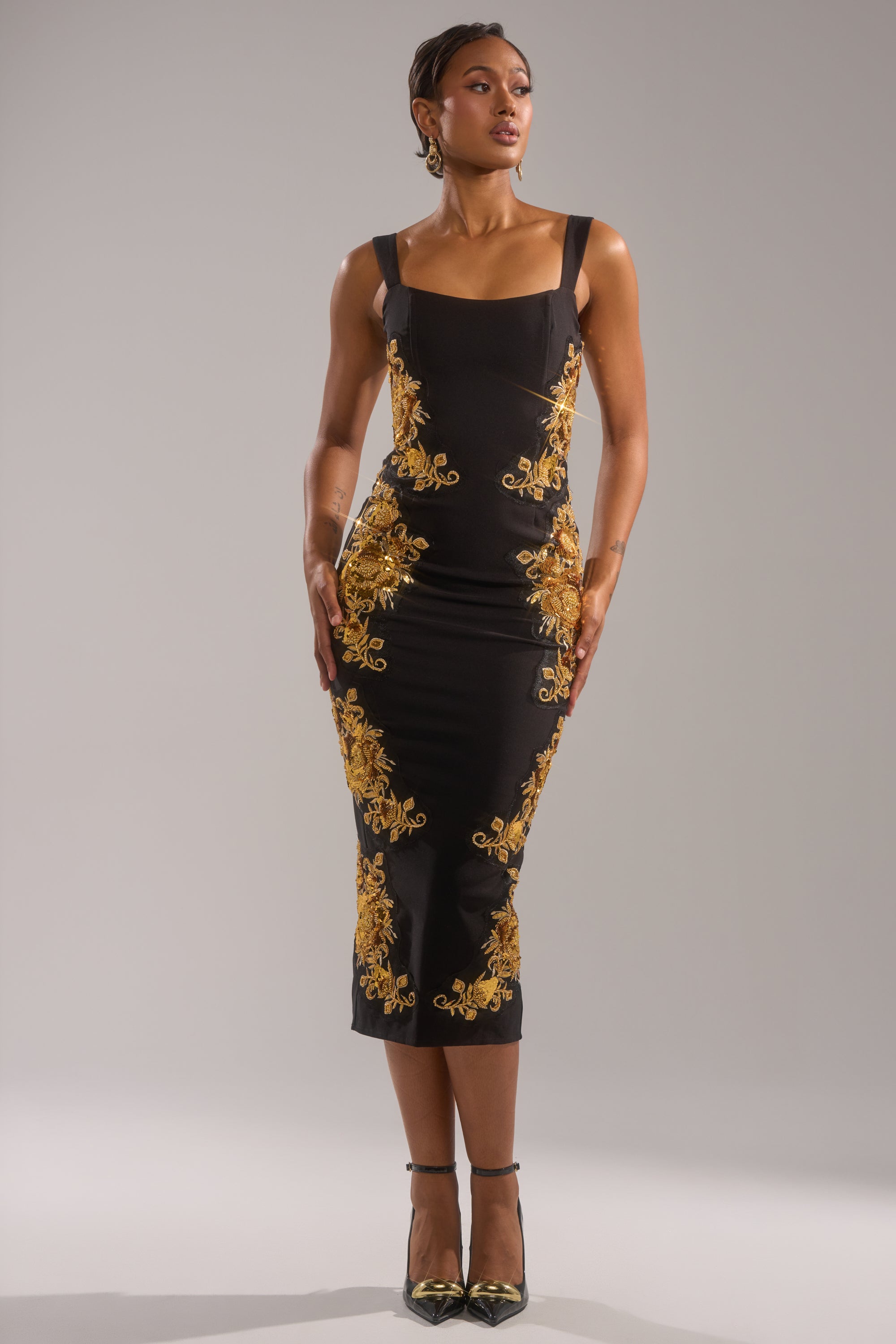 A woman stands confidently in a fitted, sleeveless black PARISIAN FLORAL EMBROIDERED MAXI DRESS with gold floral designs, paired with black heels and gold earrings against a plain gray background.