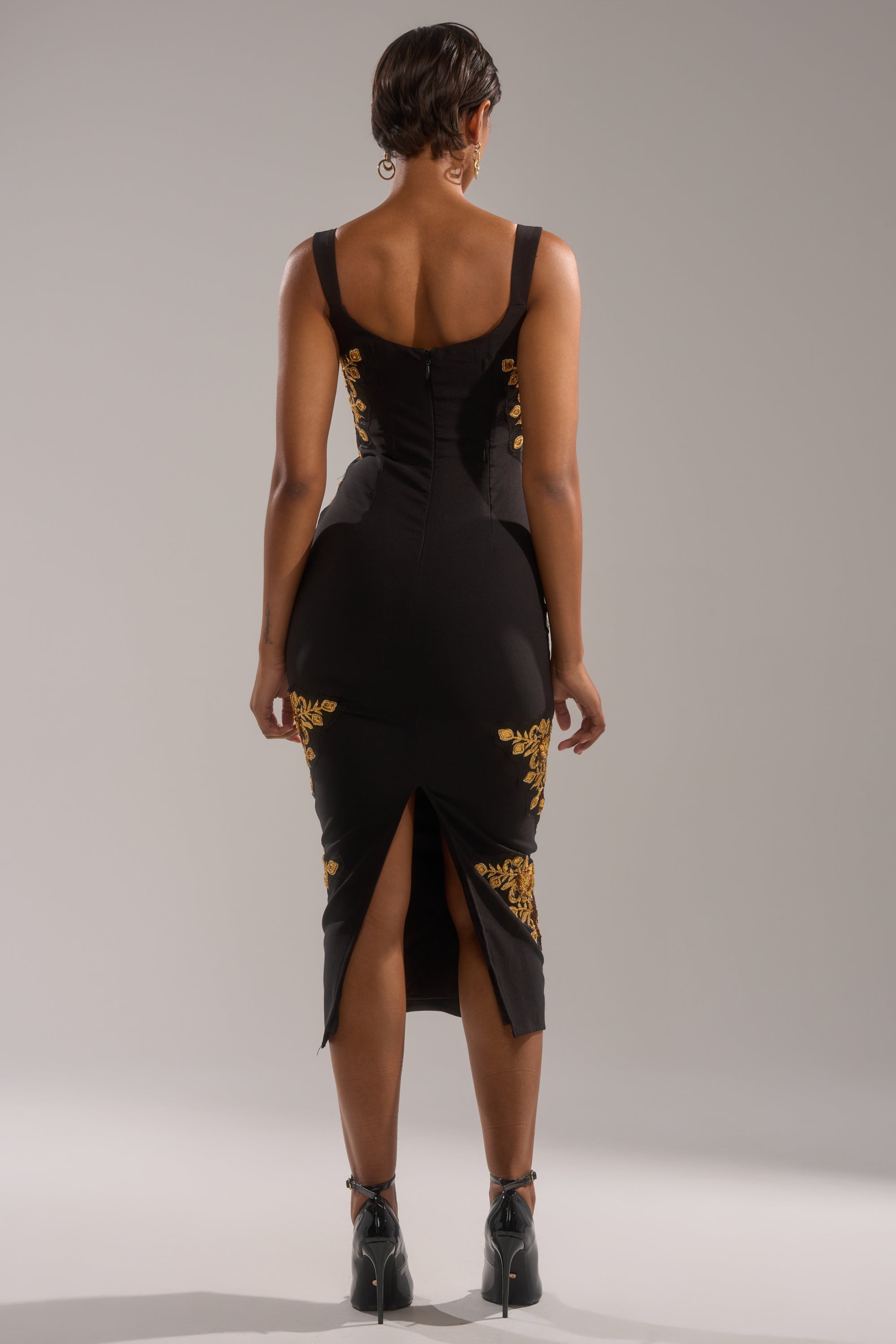 A woman with short hair faces away, wearing the PARISIAN FLORAL EMBROIDERED MAXI DRESS featuring gold embroidery and a back slit. She stands on a plain background, paired with black high heels.