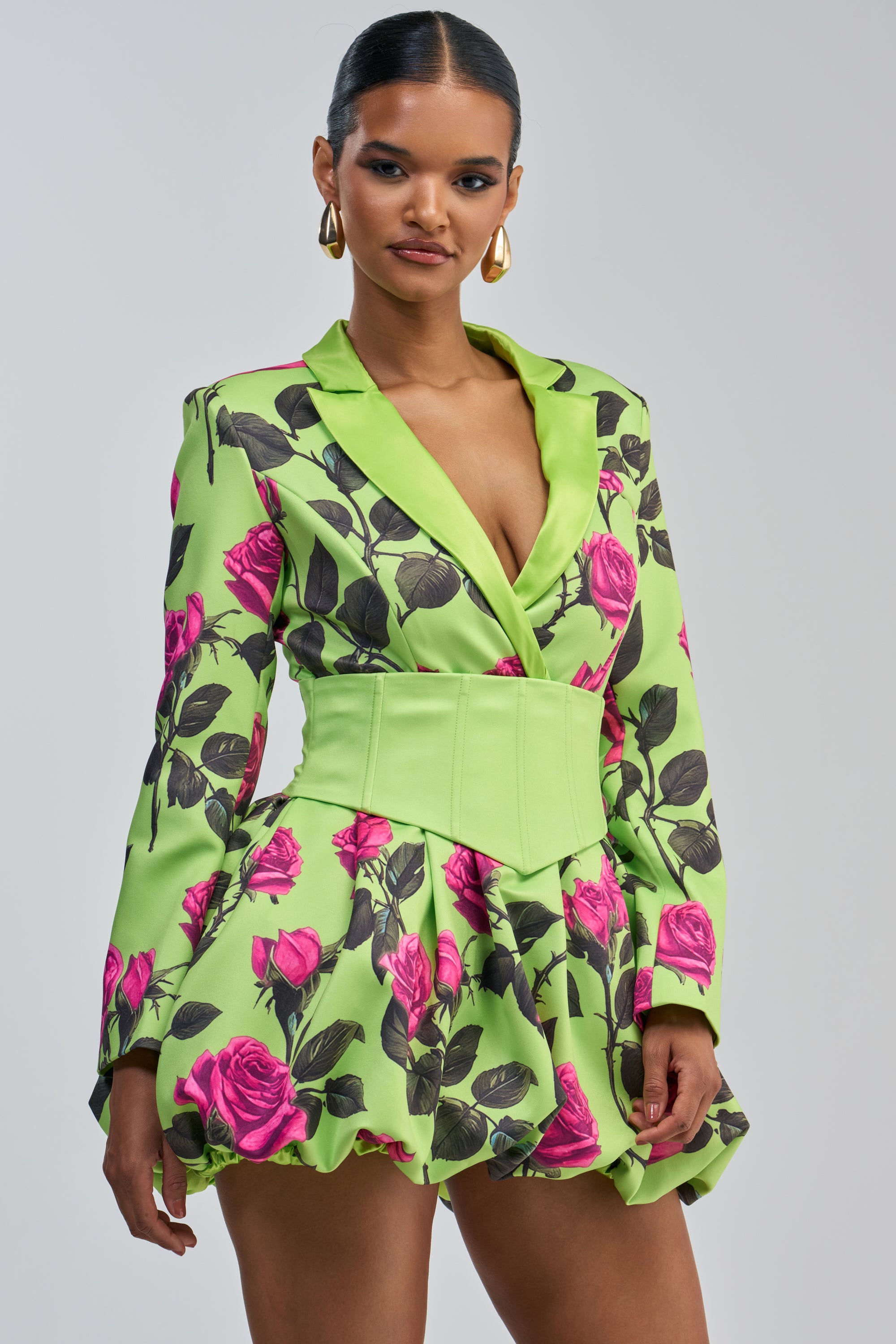 A woman wears the PAINTING THE ROSES GREEN BUBBLE MINI DRESS WITH CORSET, which features a rose graphic design in a green blazer style with pink rose patterns and a wide light green belt. She accessorizes with gold hoop earrings and has her hair slicked back, standing against a plain light gray background.