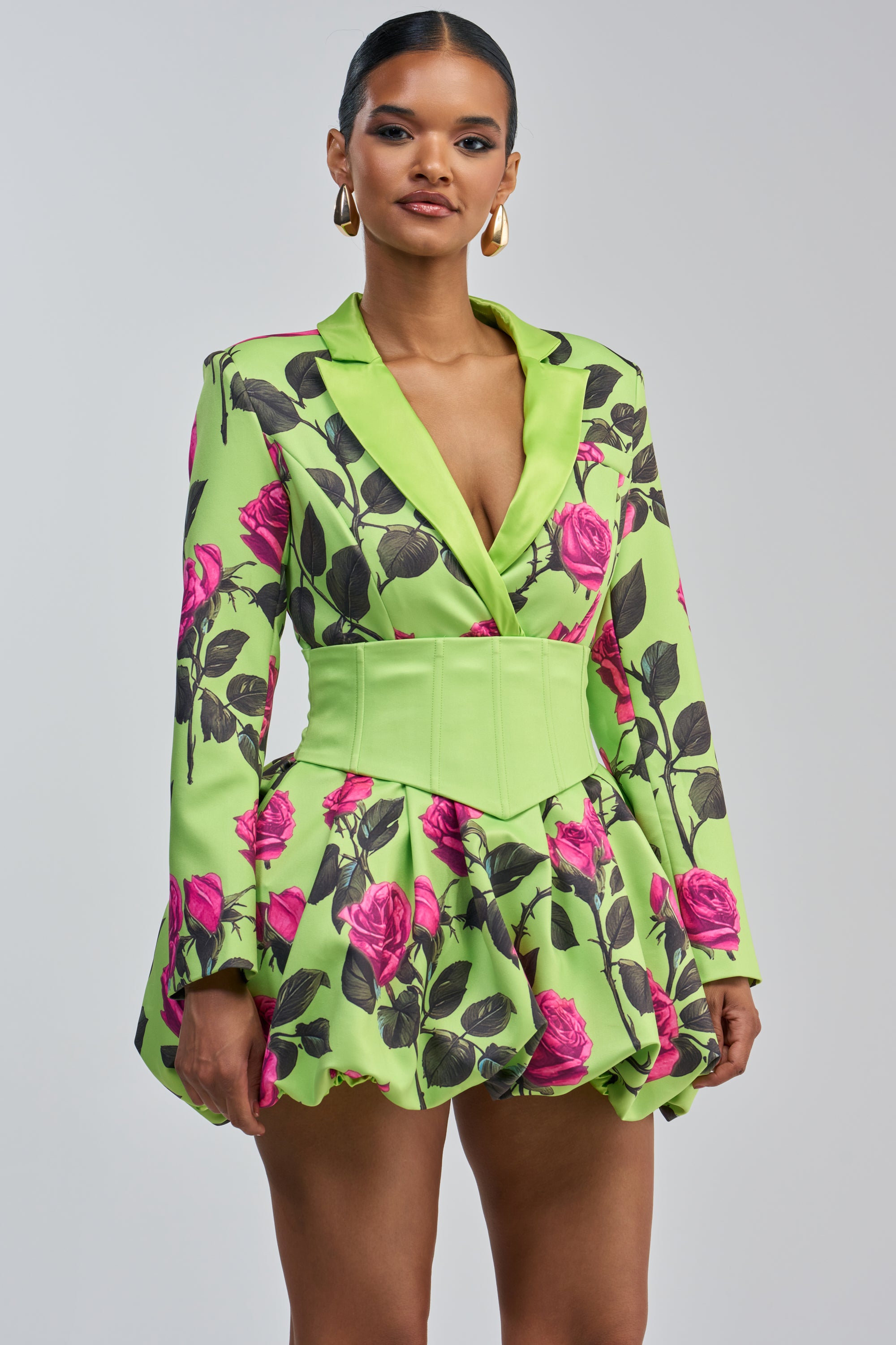 A woman stands against a plain light gray background, wearing the PAINTING THE ROSES GREEN BUBBLE MINI DRESS WITH CORSET—a lime green blazer-inspired mini dress featuring a vibrant pink rose print, flared skirt, and wide matching belt. She accessorizes with gold hoop earrings and has her hair slicked back.