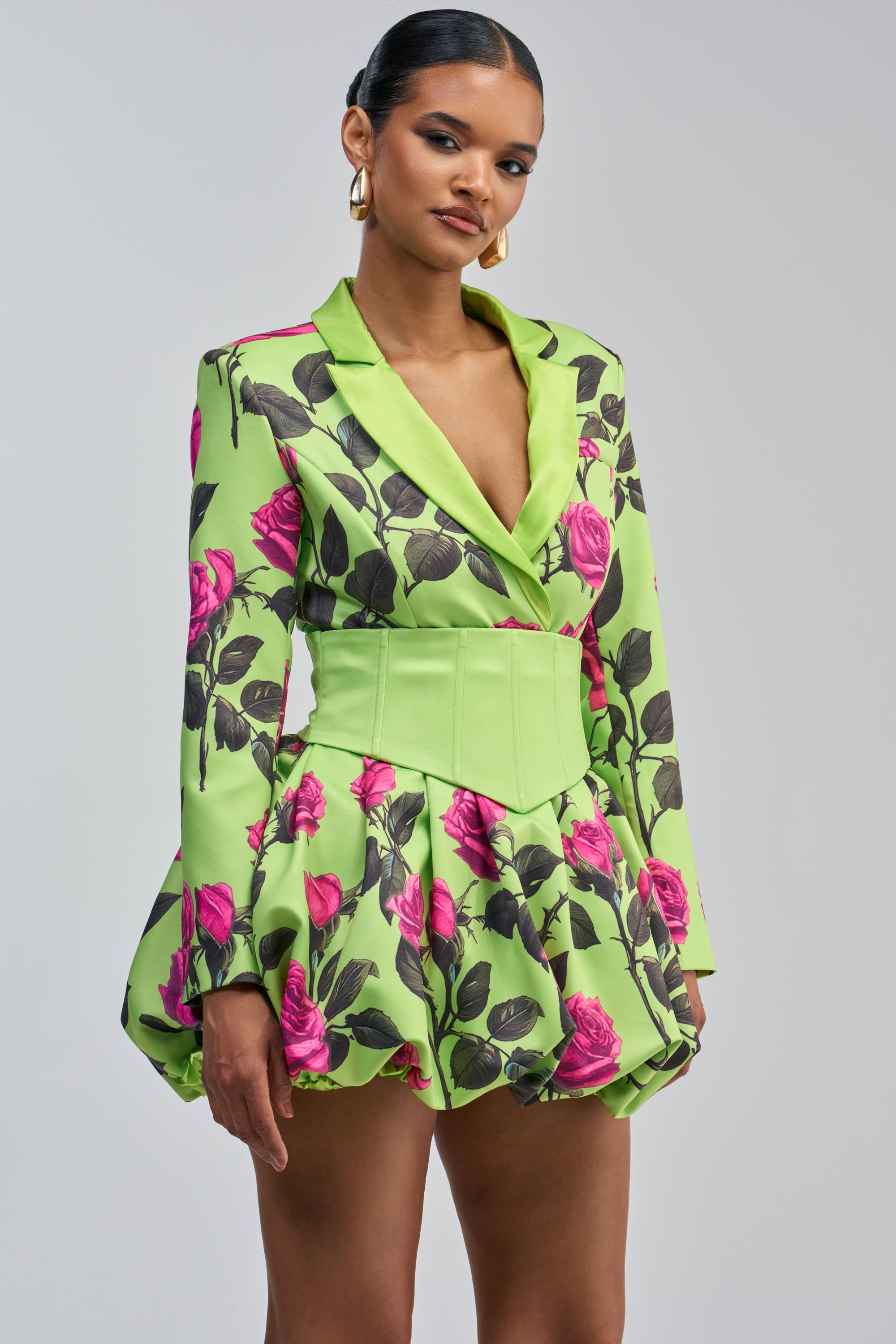 A woman wearing the PAINTING THE ROSES GREEN BUBBLE MINI DRESS WITH CORSET, a green corseted mini dress with a floral blazer-style design featuring pink roses, a wide matching belt, and a puffed skirt. She accessorizes with gold hoop earrings and has her hair styled back.