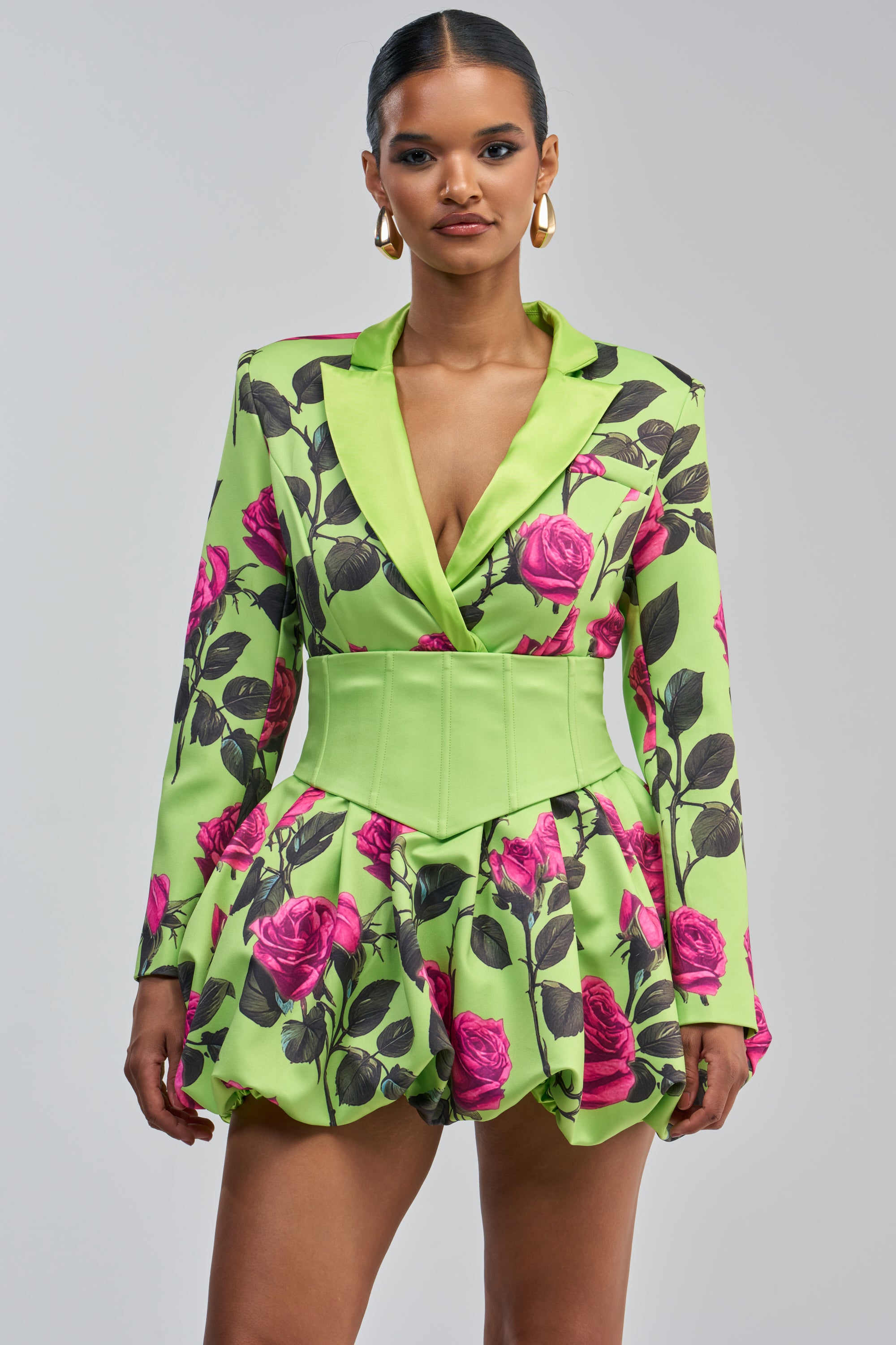 A woman wears the PAINTING THE ROSES GREEN BUBBLE MINI DRESS WITH CORSET, styled as a green blazer dress adorned with pink rose prints. The look features a puffy skirt, structured light green waistband, gold hoop earrings, and slicked-back hair, standing against a plain gray background.