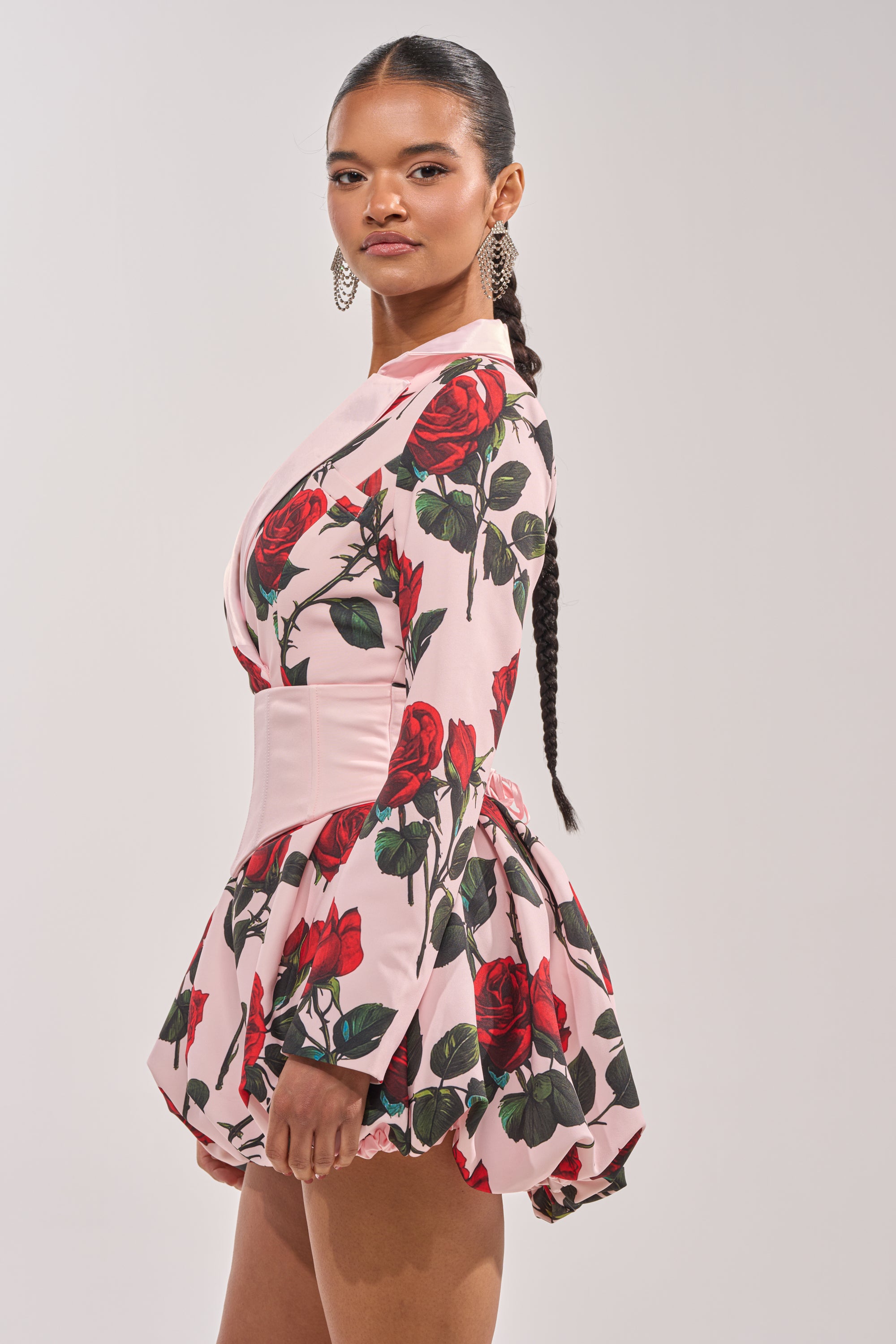 A woman with long braided hair models the PAINTING THE ROSES RED BUBBLE MINI DRESS WITH CORSET, featuring rose prints, long sleeves, and a wide light pink waistband. She stands sideways against a plain background, looking confidently at the camera. 2
