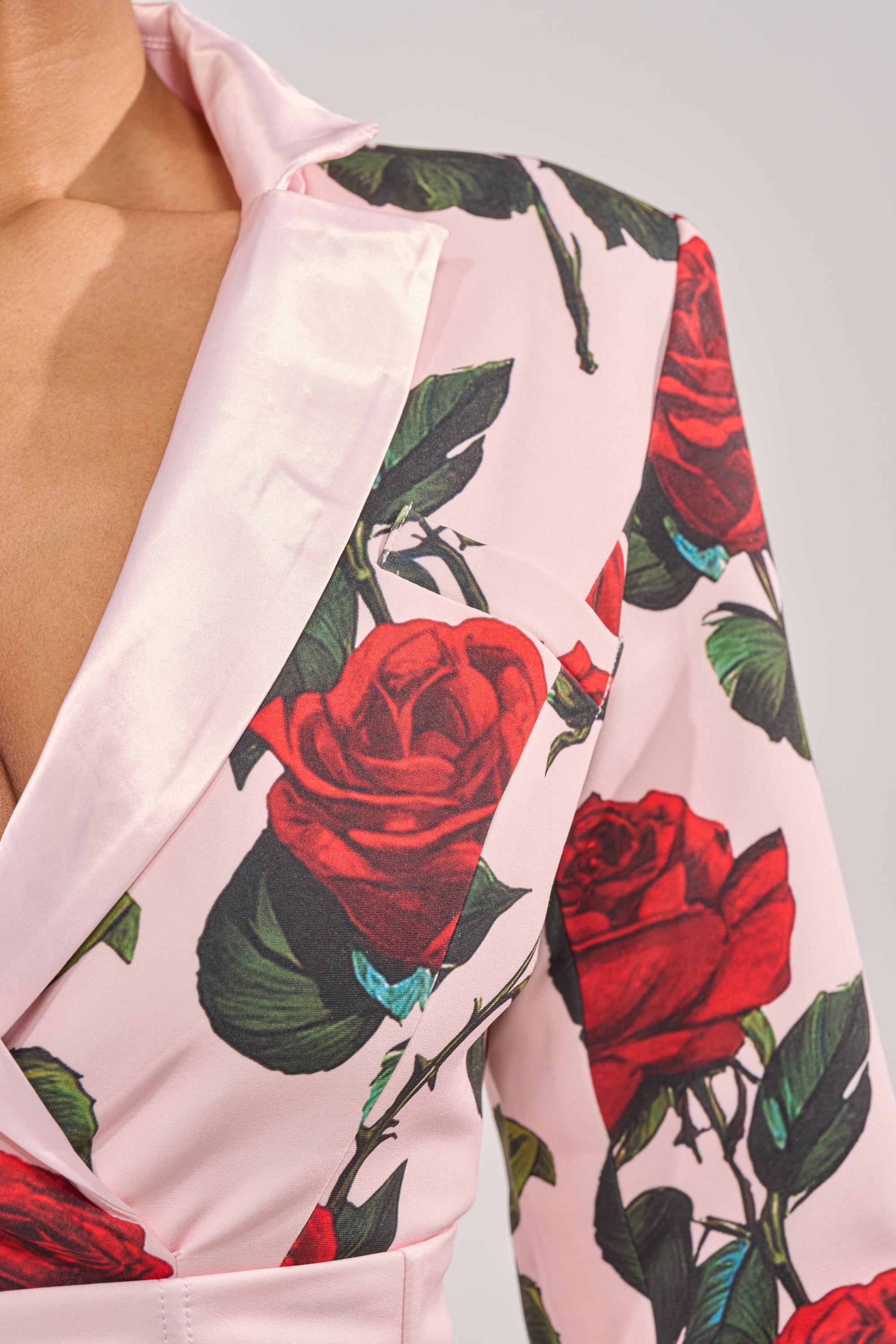 A close-up of a person wearing the PAINTING THE ROSES RED BUBBLE MINI DRESS WITH CORSET, featuring a light pink floral print with bold red roses and green leaves, notched lapel, and a partially open chest design. 6