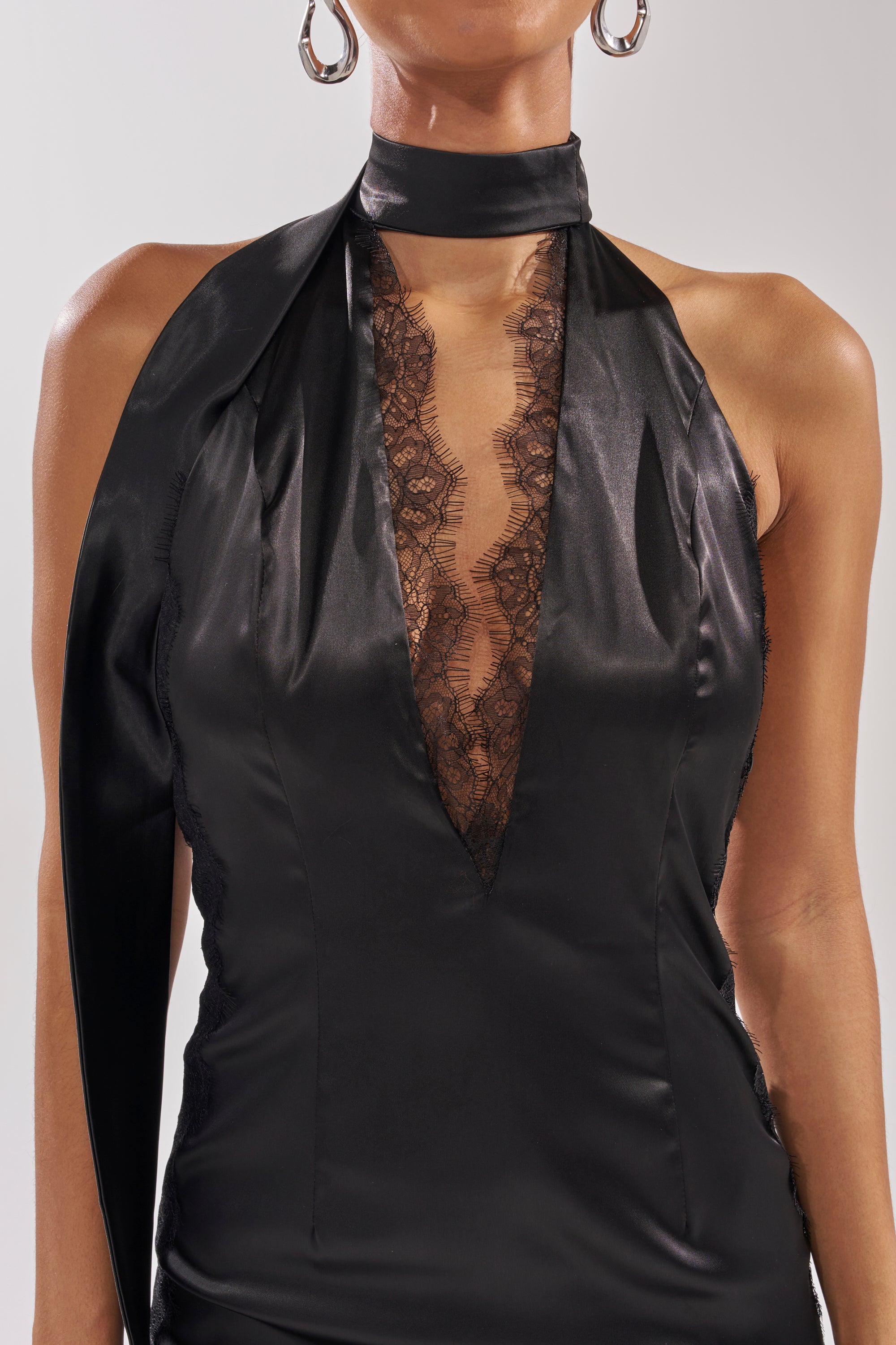 A woman wears the ALEXA SATIN MINI DRESS, a sleeveless satin mini dress with a deep V-neck lined with lace trim and a high neck collar, paired with large hoop earrings.