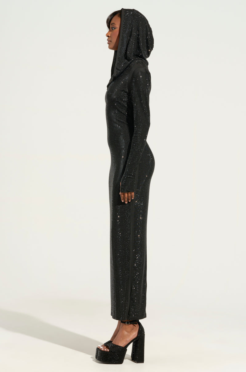 Standing against a plain light background, a person in profile wears the IMA SHINE RHINESTONE HOODED MIDI DRESS IN BLACK. The sparkling black dress, featuring long sleeves and a fitted design, is paired with high-heeled shoes.