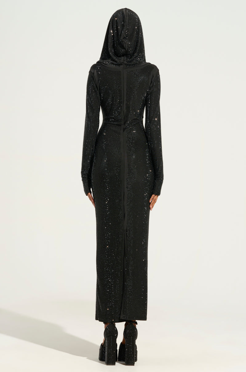 A person stands with their back to the camera, dressed in the IMA SHINE RHINESTONE HOODED MIDI DRESS IN BLACK. The dress, which is embellished with black sequins, fits snuggly and shimmers under the light. It is paired with high platform shoes against a plain, light-colored background.