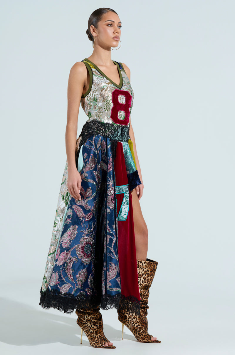 A woman exudes confidence in a CLEAT CHASER layered jersey high-low dress enhanced with floral brocade and a bold red number "8," paired with tall leopard-print boots and slicked-back hair.
