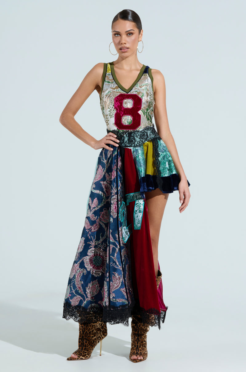 A woman confidently poses in the CLEAT CHASER LAYERED JERSEY HIGH LOW DRESS IN BROCADE, accessorizing with hoop earrings and leopard print ankle boots against a light background, showcasing the dress's vibrant charm.