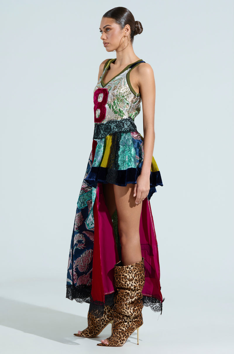 A model stands in profile wearing the CLEAT CHASER LAYERED JERSEY HIGH LOW DRESS IN BROCADE, featuring a large red "8" amid its brocade patterns. She pairs it with high leopard print boots and gold hoop earrings, her hair elegantly styled in a bun.