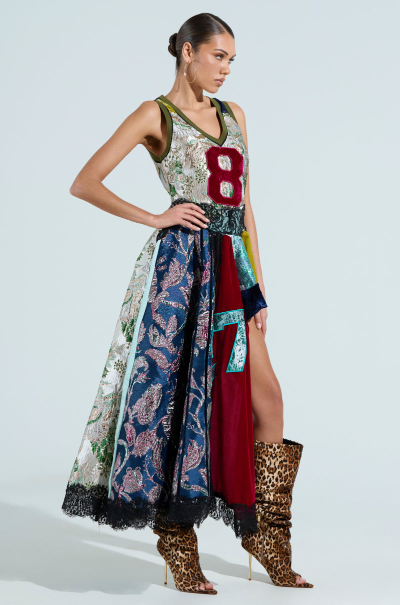 A woman poses in a CLEAT CHASER LAYERED JERSEY HIGH LOW DRESS IN BROCADE, featuring asymmetrical design with mixed patterns and a prominent "8" on the front. She pairs it with tall leopard-print boots and her hair styled in a bun, against a light backdrop.