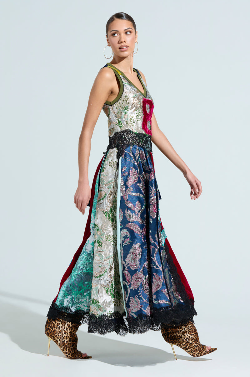 A woman poses in the CLEAT CHASER LAYERED JERSEY HIGH LOW DRESS IN BROCADE, a colorful patchwork dress with floral patterns and lace hem, featuring an asymmetrical maxi style with a burgundy number "8." She accessorizes with hoop earrings and leopard print high-heeled boots against a light backdrop.