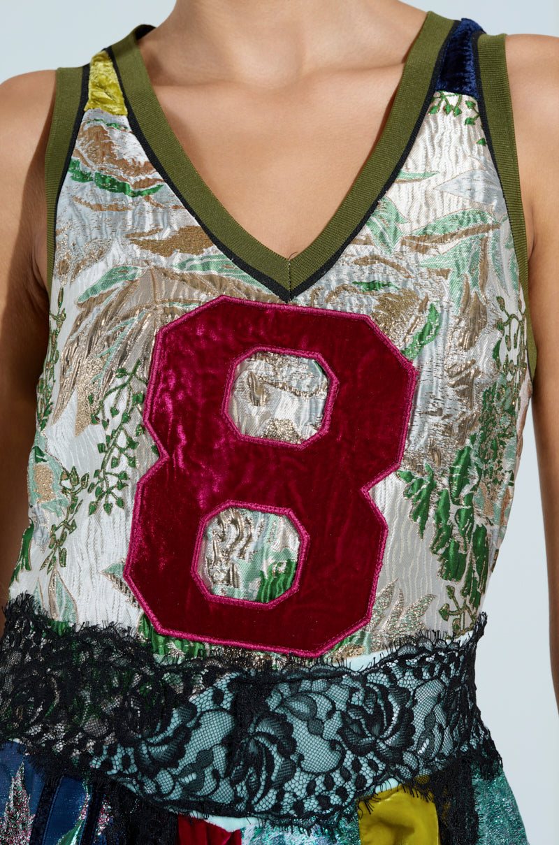 A person is wearing the CLEAT CHASER Layered Jersey High Low Dress in Brocade, featuring a textured, multicolored tank top with a green V-neckline and a large red velvet number "8" on the chest, adorned with floral patterns and lace fringe at the hem.