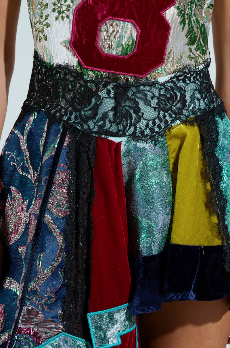 The CLEAT CHASER LAYERED JERSEY HIGH LOW DRESS IN BROCADE features a colorful patchwork skirt with velvet and metallic fabrics, a large red number eight on the bodice, and black lace trim at the waist. It includes intricate floral and abstract patterns throughout.