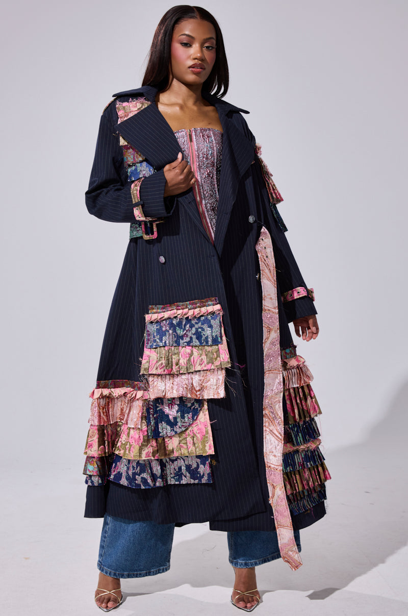 A woman stands confidently against a plain background, wearing the MAKE AN ENTRANCE PATCHWORK TRENCH with colorful, layered floral and patterned ruffles, styled with wide blue jeans and a matching bustier.