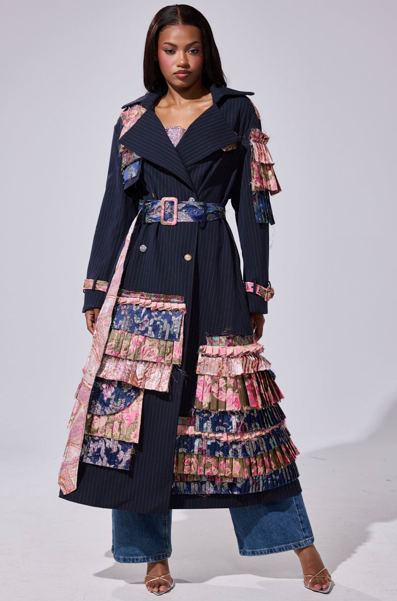 A woman stands confidently in the MAKE AN ENTRANCE PATCHWORK TRENCH, an oversized longline jacket with brocade accents and layered floral ruffles, cinched with a matching belt. She pairs it with blue jeans and wears her hair straight against a plain background.