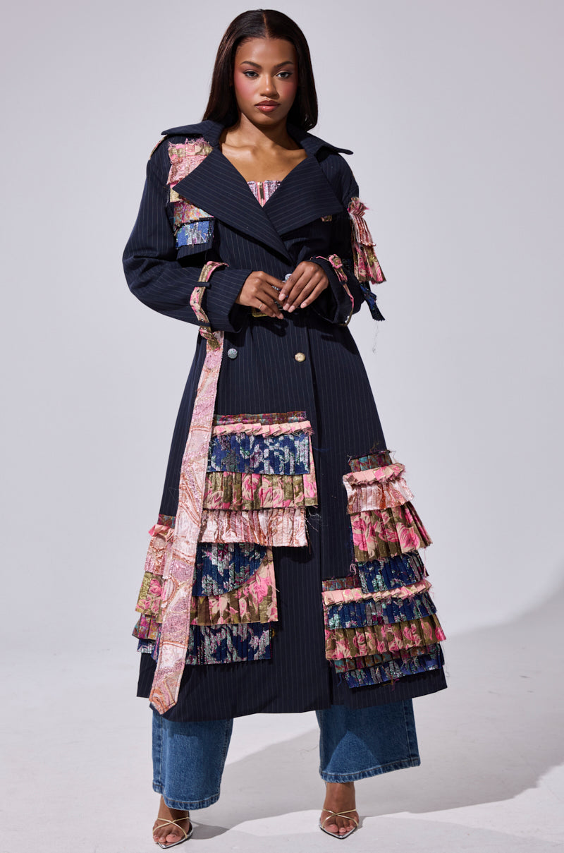 A woman models the MAKE AN ENTRANCE PATCHWORK TRENCH, featuring colorful layered ruffles and a pink patterned belt, styled with blue jeans and strappy heels against a simple light backdrop.