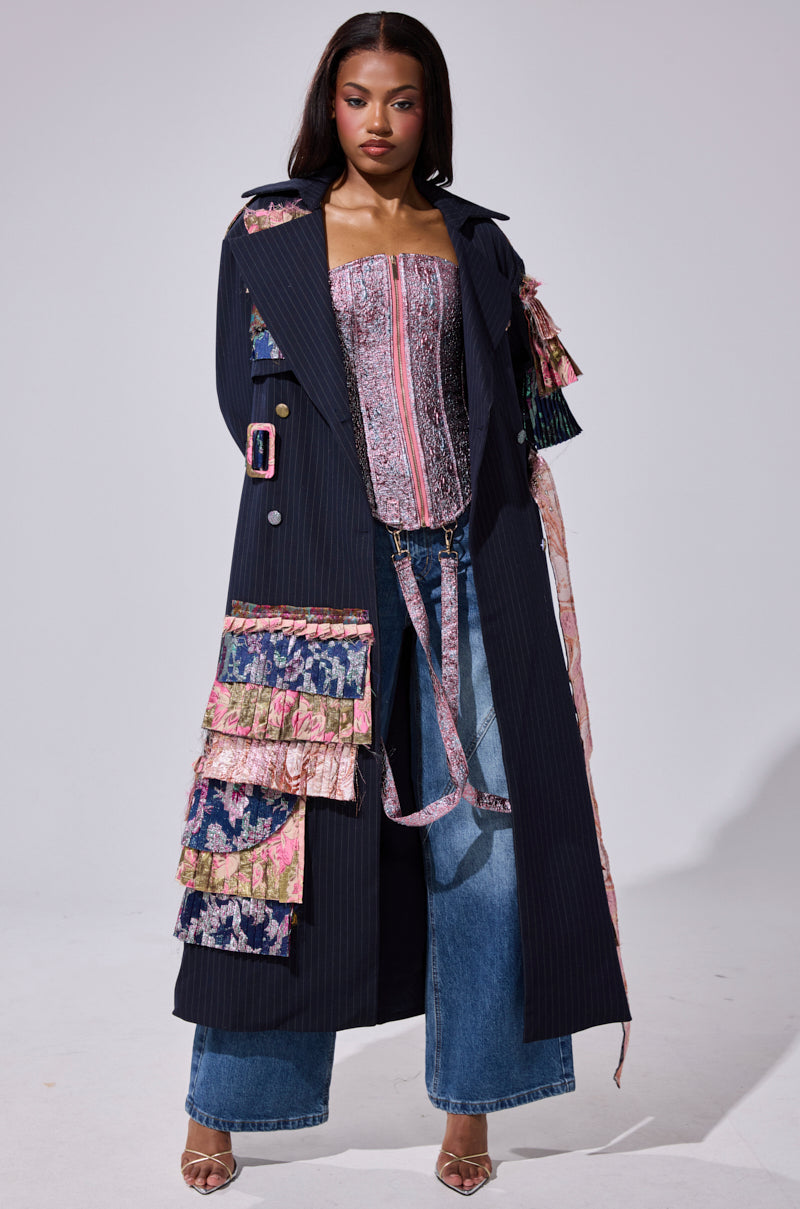 A woman wears the MAKE AN ENTRANCE PATCHWORK TRENCH over a pink and silver corset top and wide-legged blue jeans, finishing her look with open-toed heels.