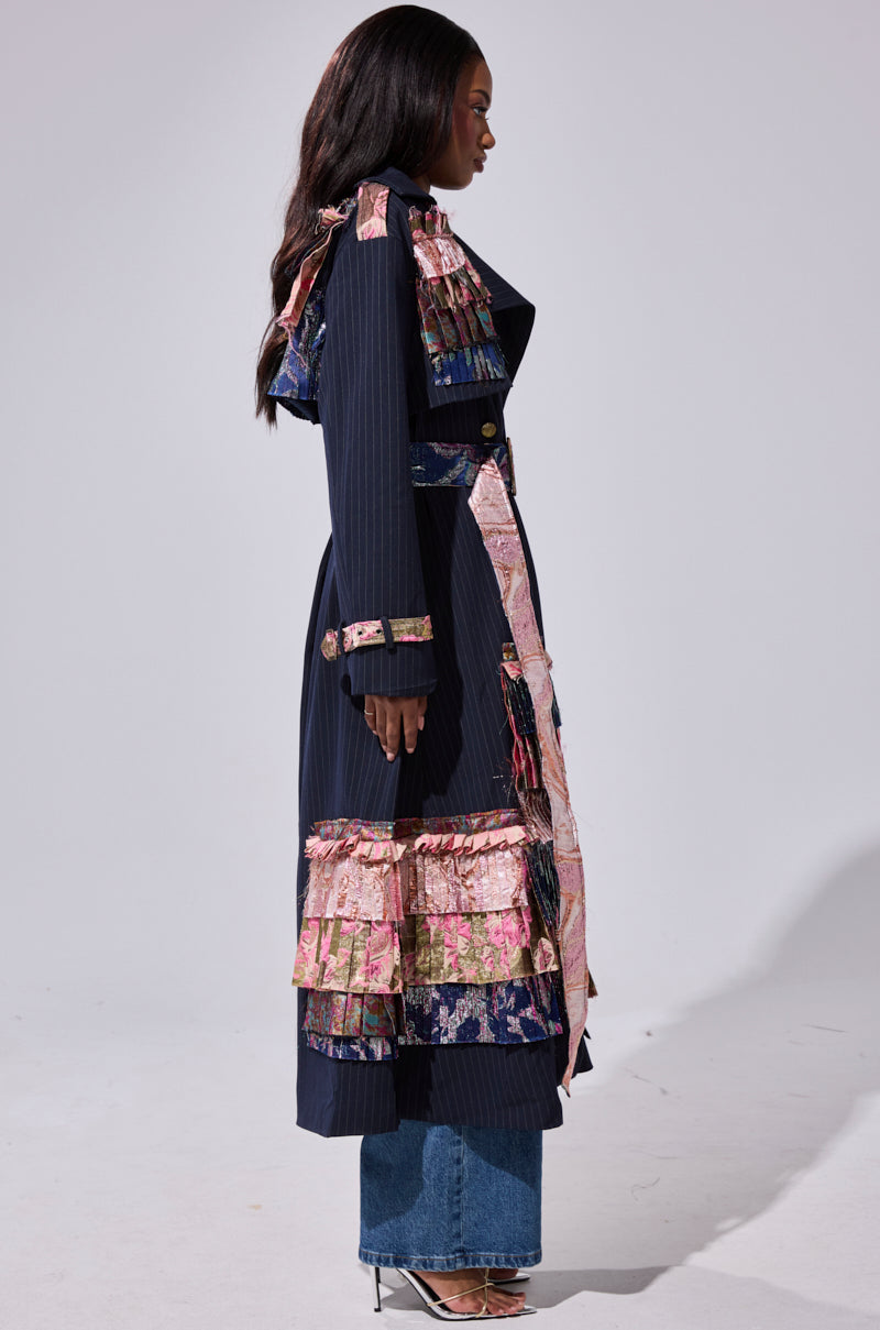 A woman stands in profile wearing the MAKE AN ENTRANCE PATCHWORK TRENCH with pink floral ruffles, styled with blue jeans and pointed heels against a plain light background.