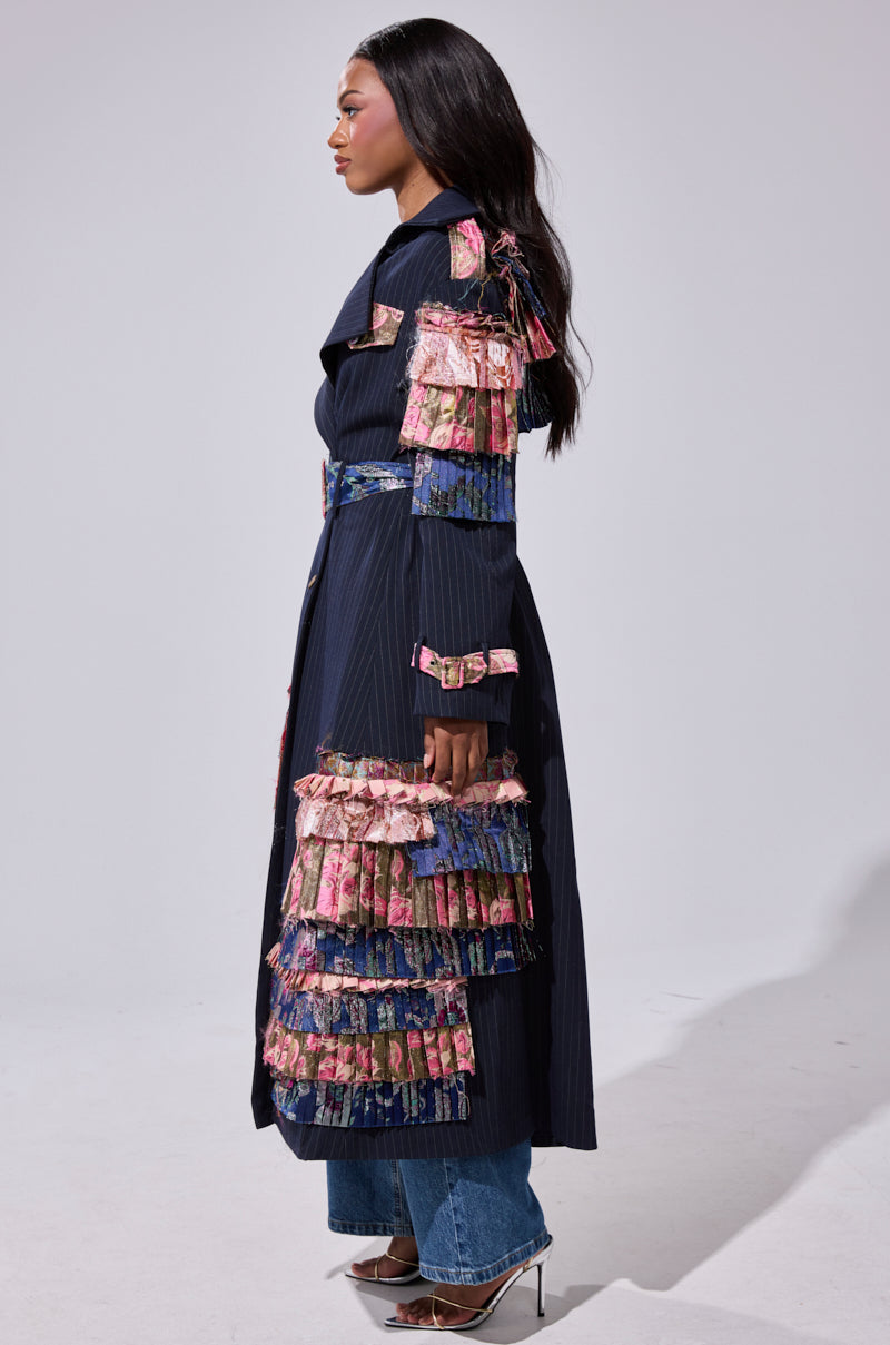 A woman stands sideways in the MAKE AN ENTRANCE PATCHWORK TRENCH, featuring layered multicolored ruffles on the sleeves and hem, paired with blue jeans and silver heeled sandals.