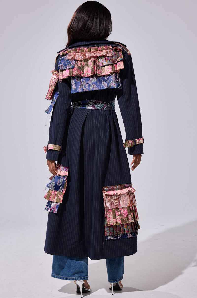 A person faces away, wearing the MAKE AN ENTRANCE PATCHWORK TRENCH, featuring colorful floral ruffles on the shoulders, cuffs, and lower back. Blue jeans and silver heeled sandals peek out from beneath the bold pinstripe coat.