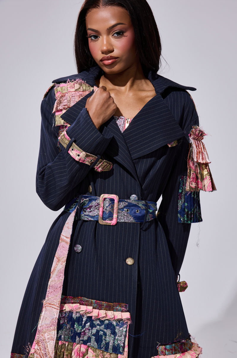 A woman models the MAKE AN ENTRANCE PATCHWORK TRENCH, an oversized longline coat with navy pinstripes, vibrant pink and floral patches, a wide collar, and matching belt. She has long dark hair and stands against a plain light background.