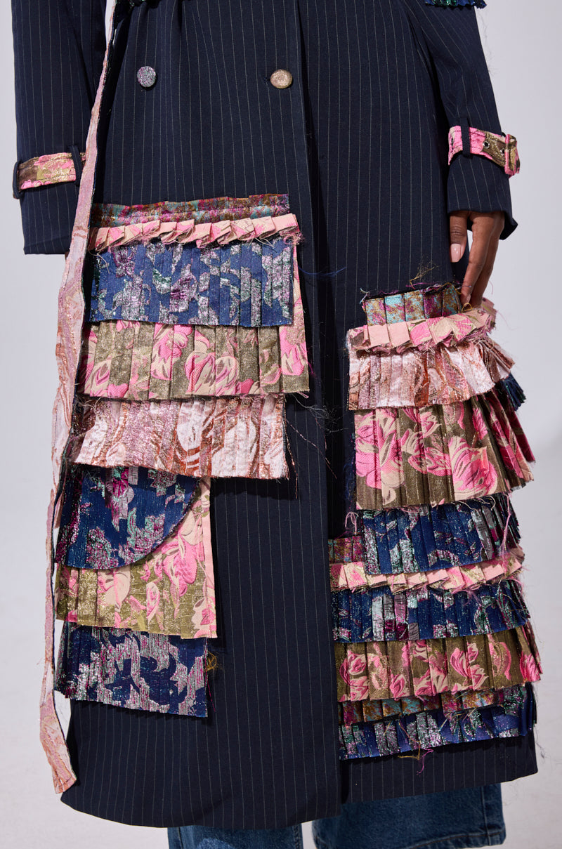 A close-up of the MAKE AN ENTRANCE PATCHWORK TRENCH, featuring navy pinstripes with layered pink, blue, and green pleated floral patches. A hand rests alongside the intricate embellishments.