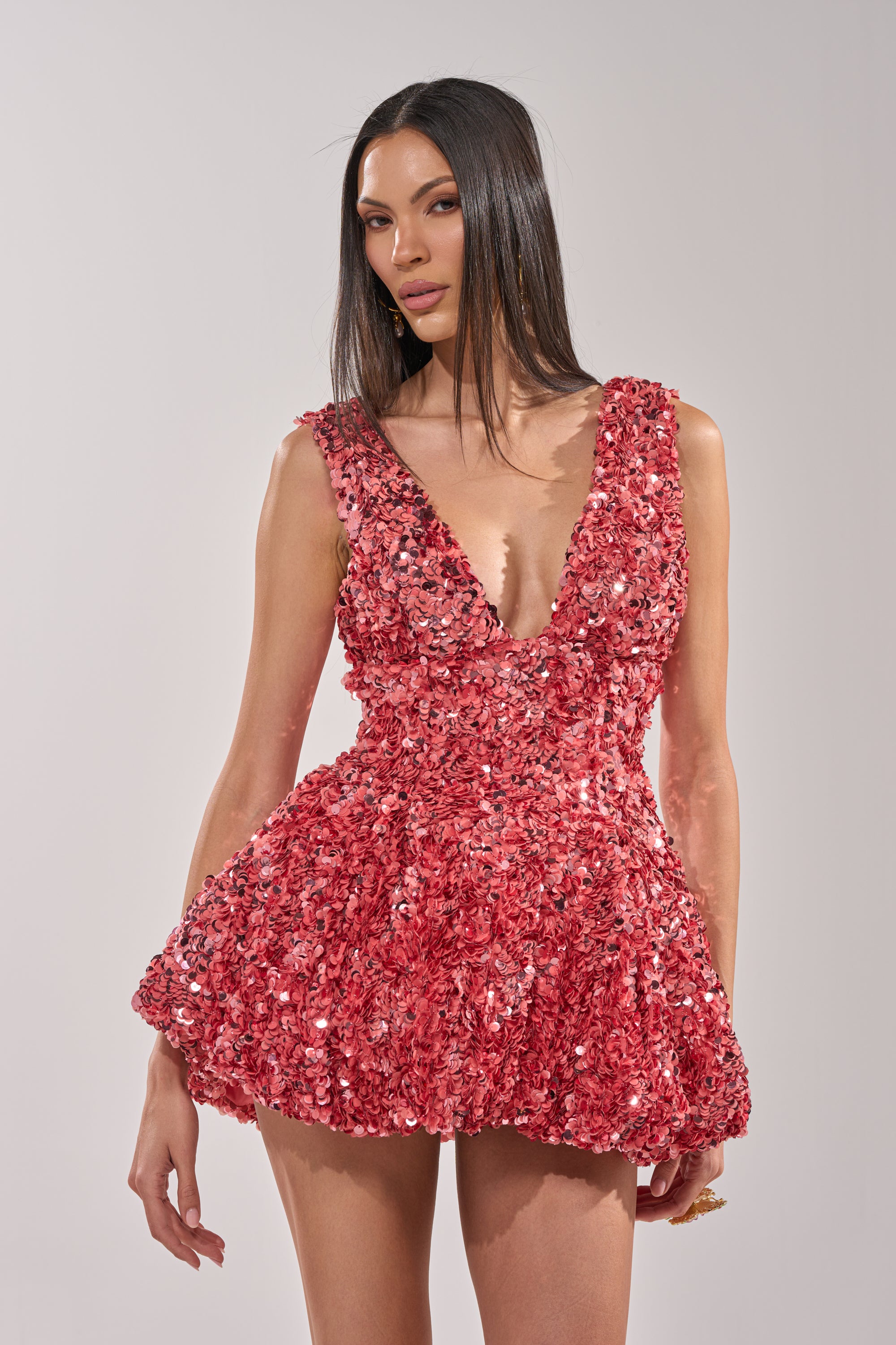 A woman with straight dark hair wears the BARBIE GIRL SEQUIN MINI DRESS, a vibrant red mini dress featuring a plunging neckline and textured floral embellishments, standing against a plain light background.