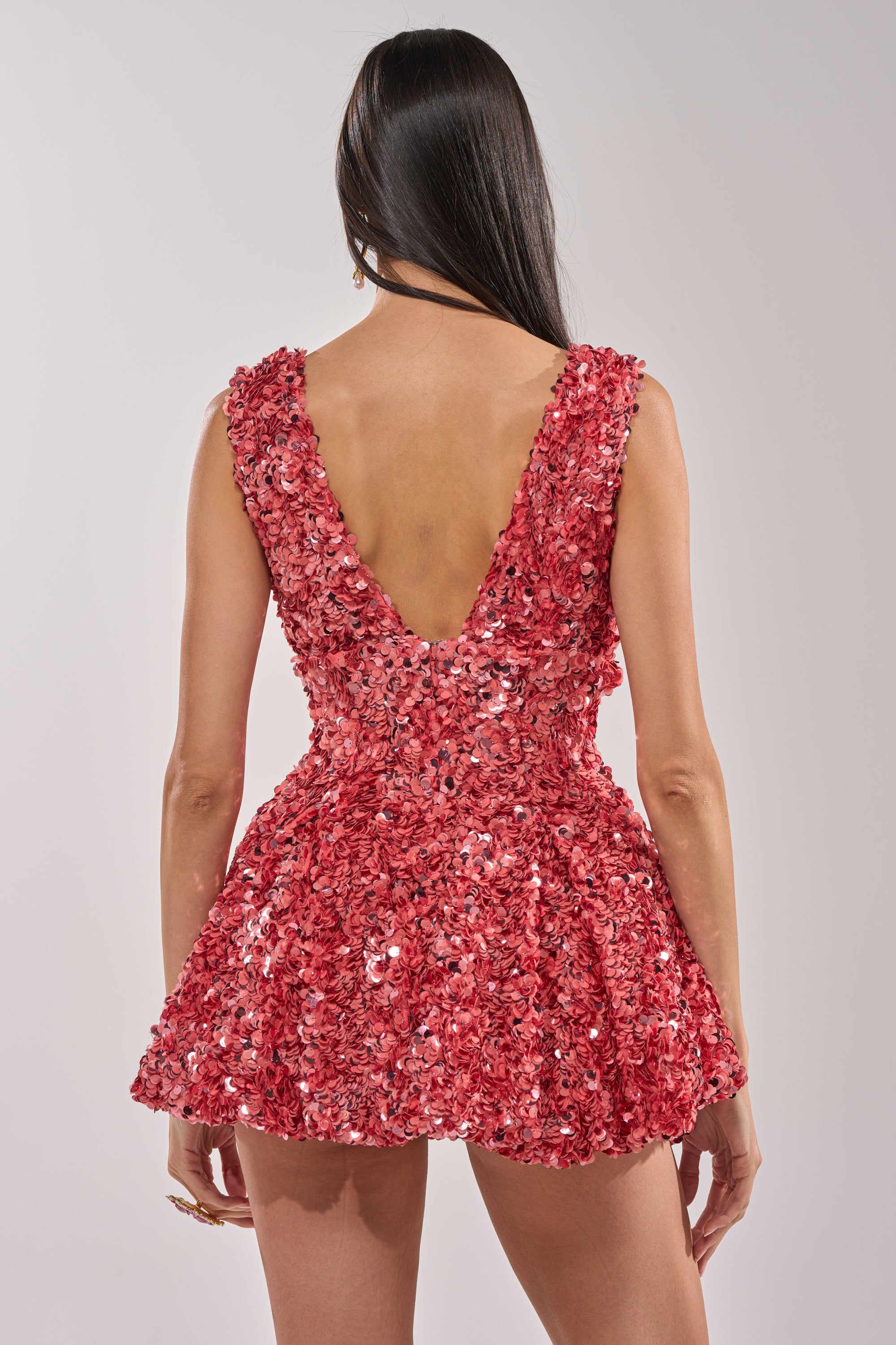 A person with long dark hair stands against a plain light background, facing away from the camera, wearing the BARBIE GIRL SEQUIN MINI DRESS in pink, featuring a sequin finish and deep V-back.