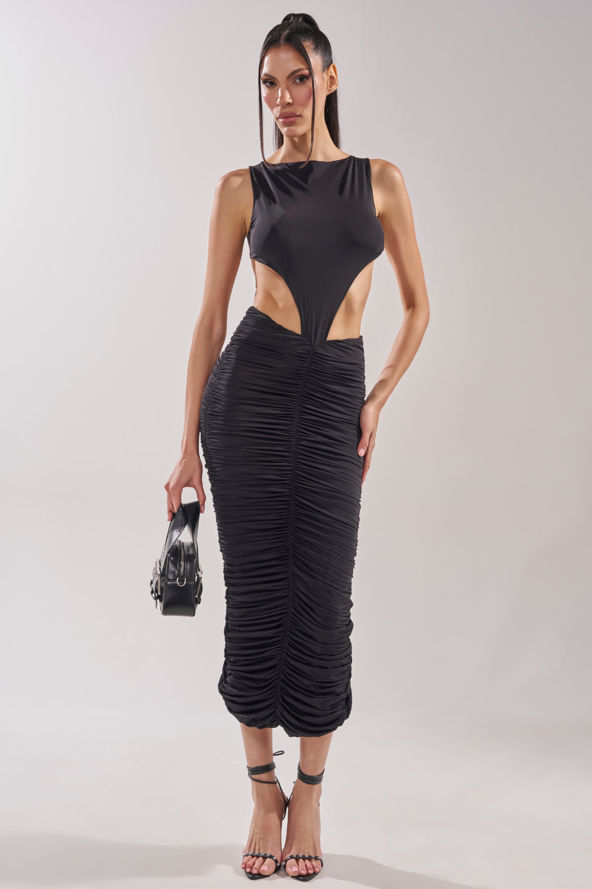 A woman models the EYES ON ME MAXI DRESS—a black cutout maxi with side cutouts and a ruched skirt. She accessorizes with a black handbag and strappy heels, her long hair styled straight against a plain, light background. 0