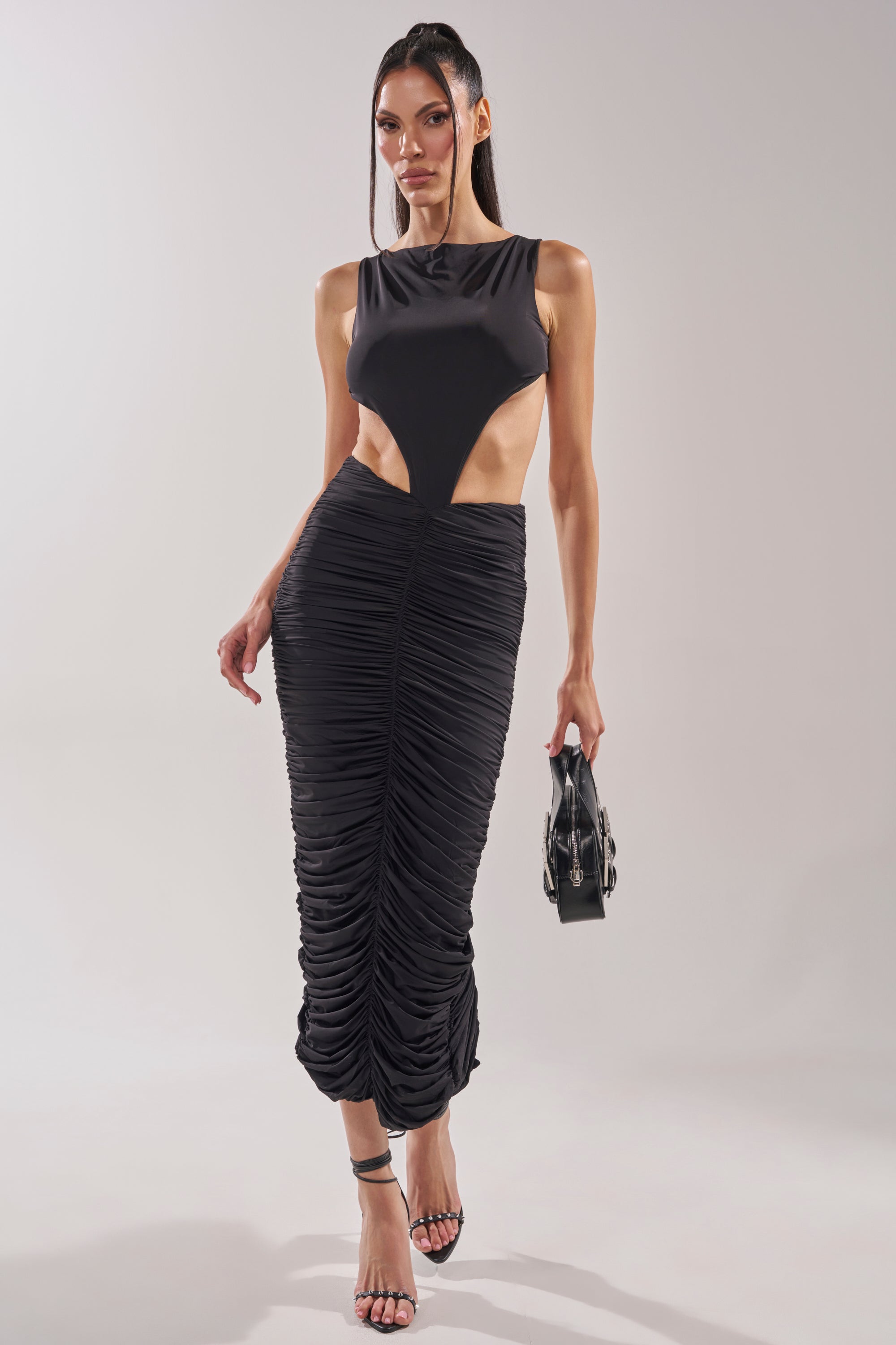 A woman wears the EYES ON ME MAXI DRESS, a black cutout maxi with a ruched skirt, paired with a small black handbag and strappy heels, standing against a plain background. 2