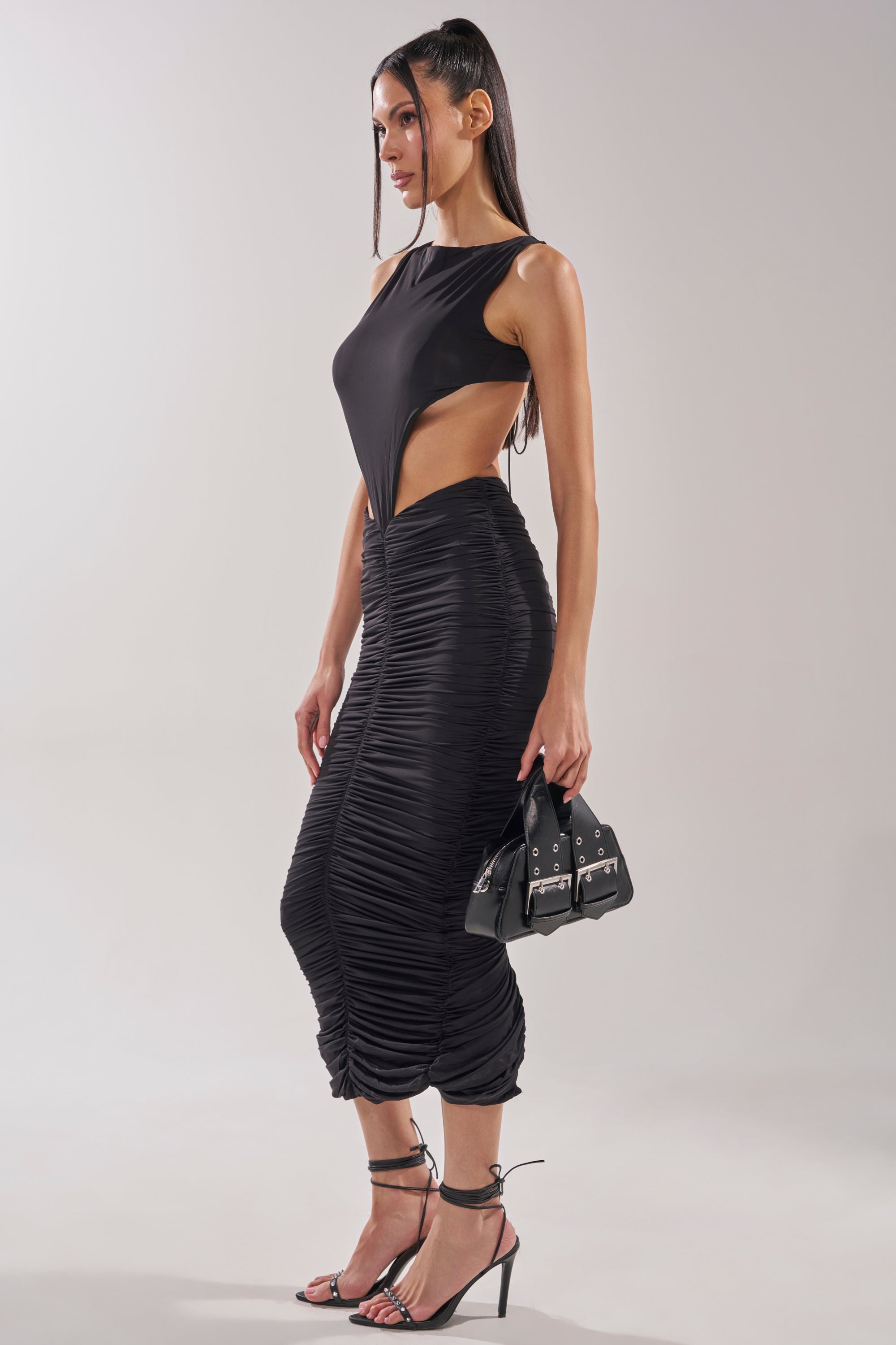 She models the EYES ON ME MAXI DRESS, a black cutout ruched maxi, paired with strappy black heels and a small black handbag. Her long, straight hair is styled sleekly against a plain light background. 3