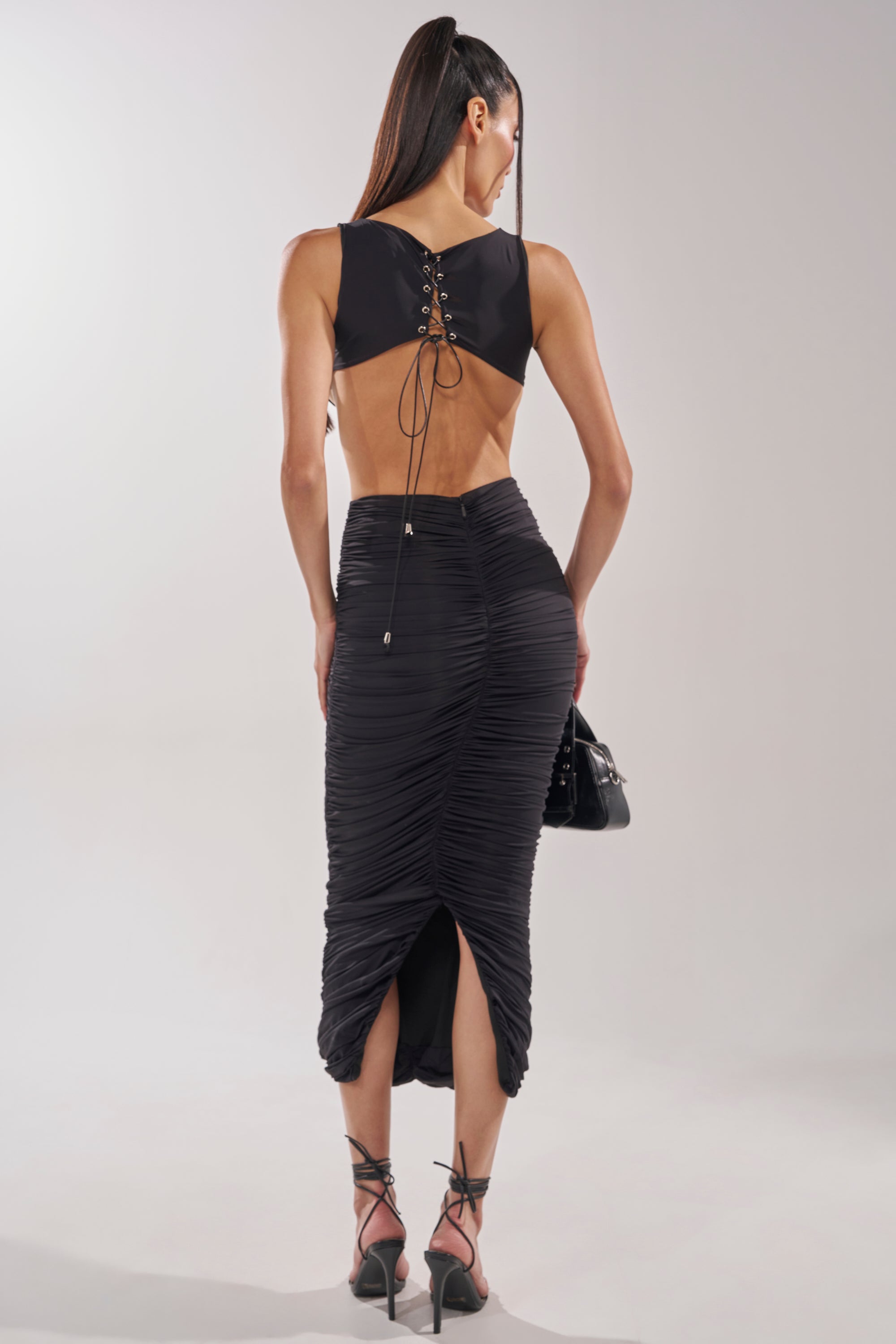 A woman with long dark hair in a high ponytail wears the EYES ON ME MAXI DRESS, a fitted black cutout style with ruched and lace-up details. She carries a black handbag and finishes her formal look with strappy high-heeled sandals. 4