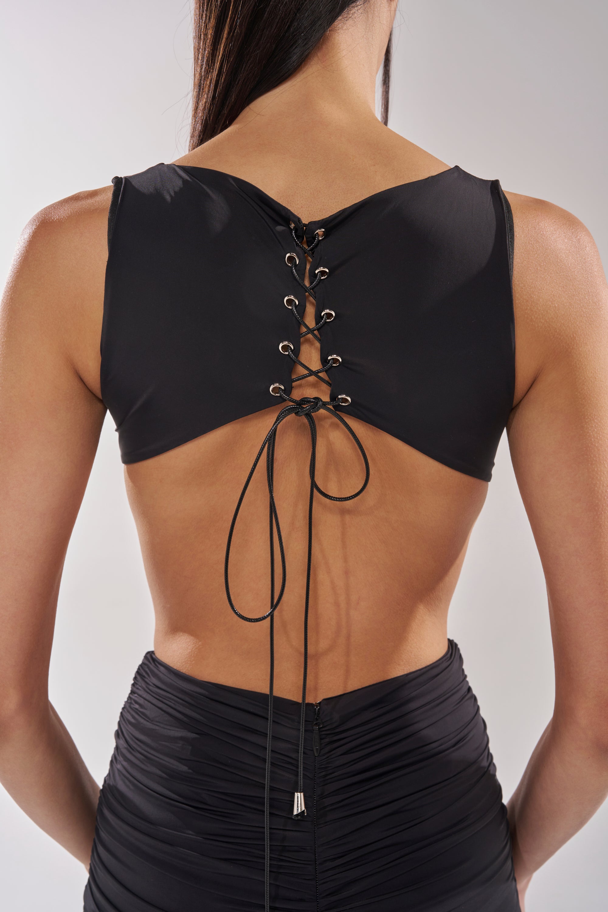 A woman is shown from behind wearing the EYES ON ME MAXI DRESS, featuring a black sleeveless design with a lace-up back, tied bow detail, and high-waisted ruched bodycon fit for an elegant modern look. 7