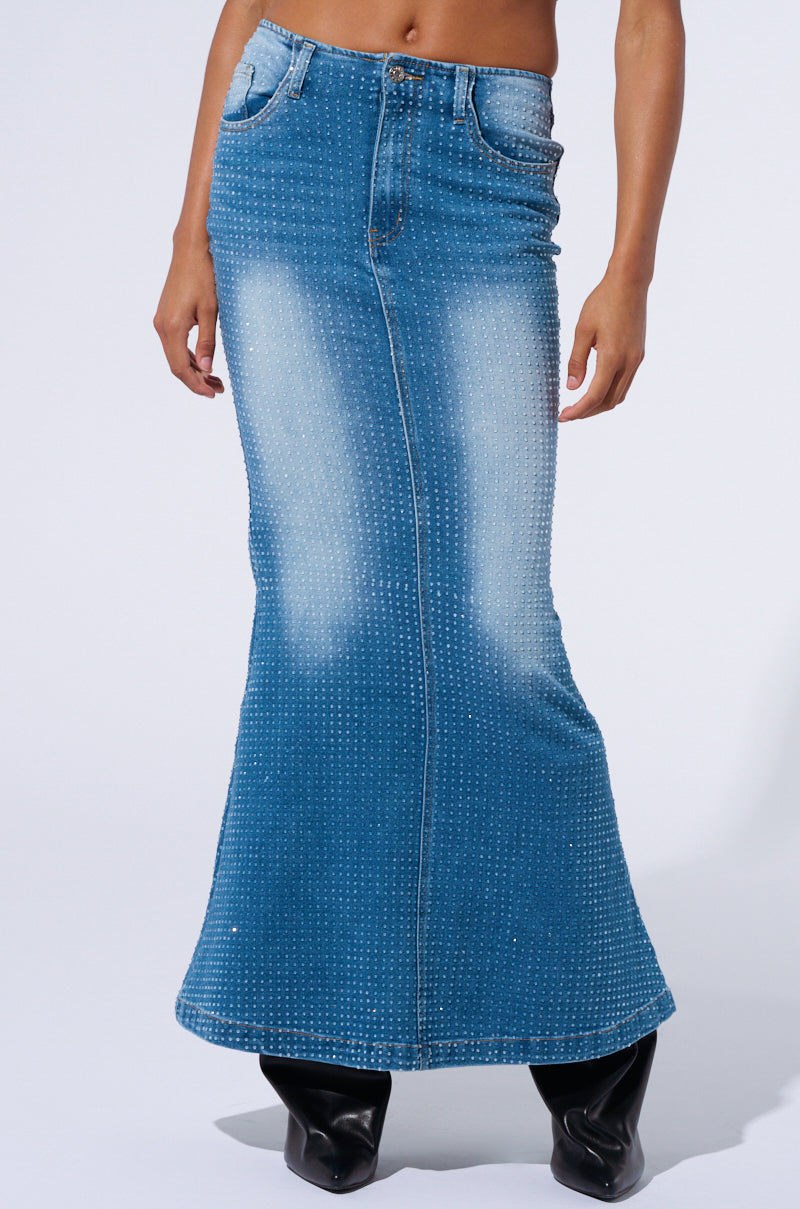 A person modeling the RICH GIRL ENERGY EMBELLISHED DENIM MAXI SKIRT adorned with tiny silver studs. This full-length denim skirt features a slight flare at the hem, faded sections along the front, and five-pocket styling. The model is also wearing black heeled boots.