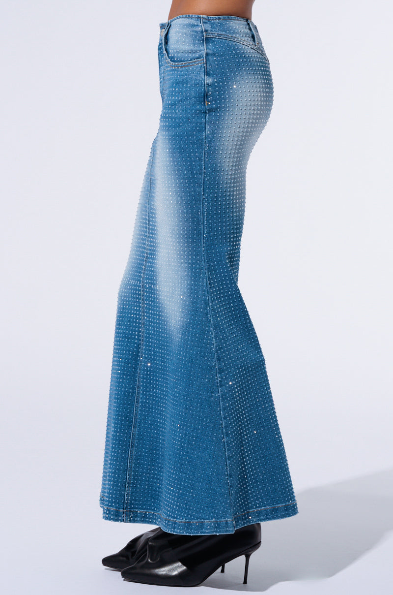 A person wearing the RICH GIRL ENERGY EMBELLISHED DENIM MAXI SKIRT stands in profile view. They are also sporting black, pointed-toe shoes with a small heel. The background is plain white.