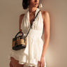 A woman showcases the SUMMER'S AROUND THE CORNER EYELET MINI DRESS—a sleeveless white mini dress with ruffles—while holding a small brown and black crossbody bag against a beige backdrop for an effortless summer look.