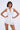 A woman with chin-length dark hair and dangling earrings poses against a plain light background, wearing the SUMMER'S AROUND THE CORNER EYELET MINI DRESS—a sleeveless white eyelet halter mini dress with a tiered skirt, perfect for summer.