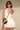 Wearing the SUMMER'S AROUND THE CORNER EYELET MINI DRESS, a woman accessorizes with sunglasses, gold earrings, and a small black and tan handbag as she poses against a beige background—a perfect summer look.